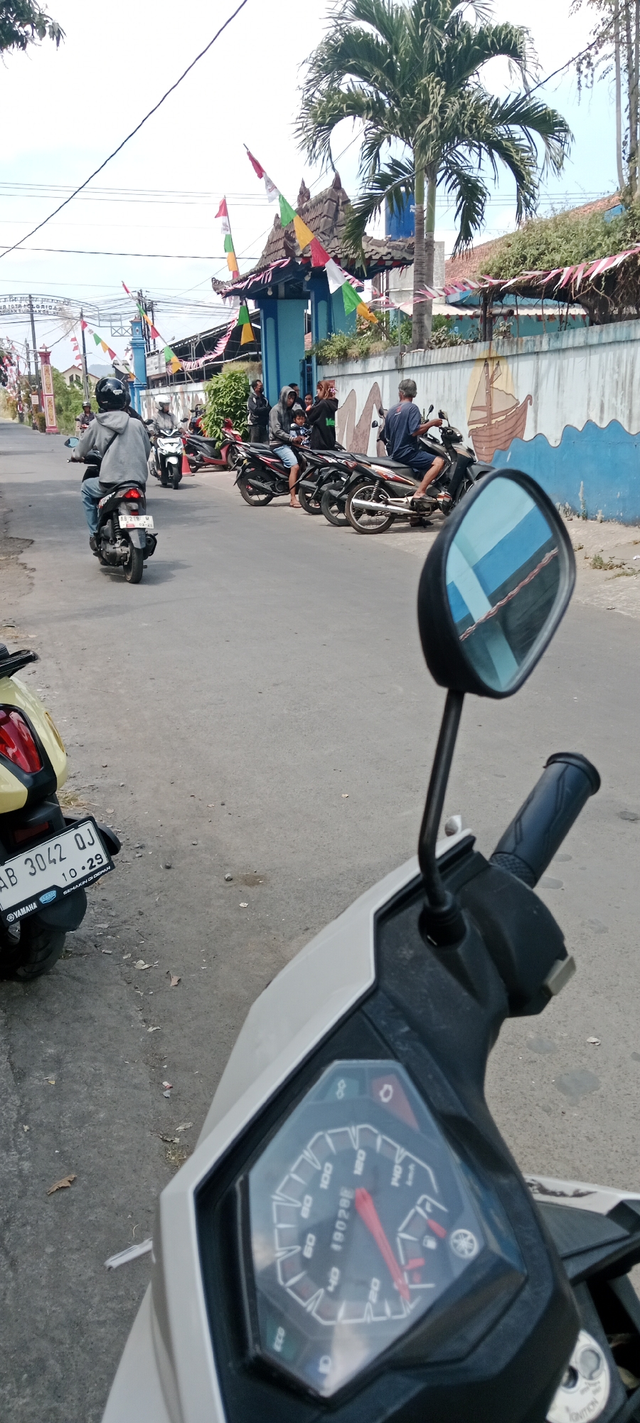 In Purwomartani, Indonesia, on a paved street lined with buildings decorated with colorful flags, several motorcycles are parked and in motion. In the foreground, a motorcycle's dashboard and side mirror are visible, reflecting the street and a palm tree. To the left, a yellow motorcycle with a license plate "AB 3042 QJ" is parked. Further down the street, a person on a grey motorcycle rides away from the camera, followed by other motorcycles and individuals near a blue building with a decorative roof. A wall with a painted mural of a ship and waves runs along the right side of the street. The sky is overcast, and the overall atmosphere suggests a daytime scene on a moderately busy road.