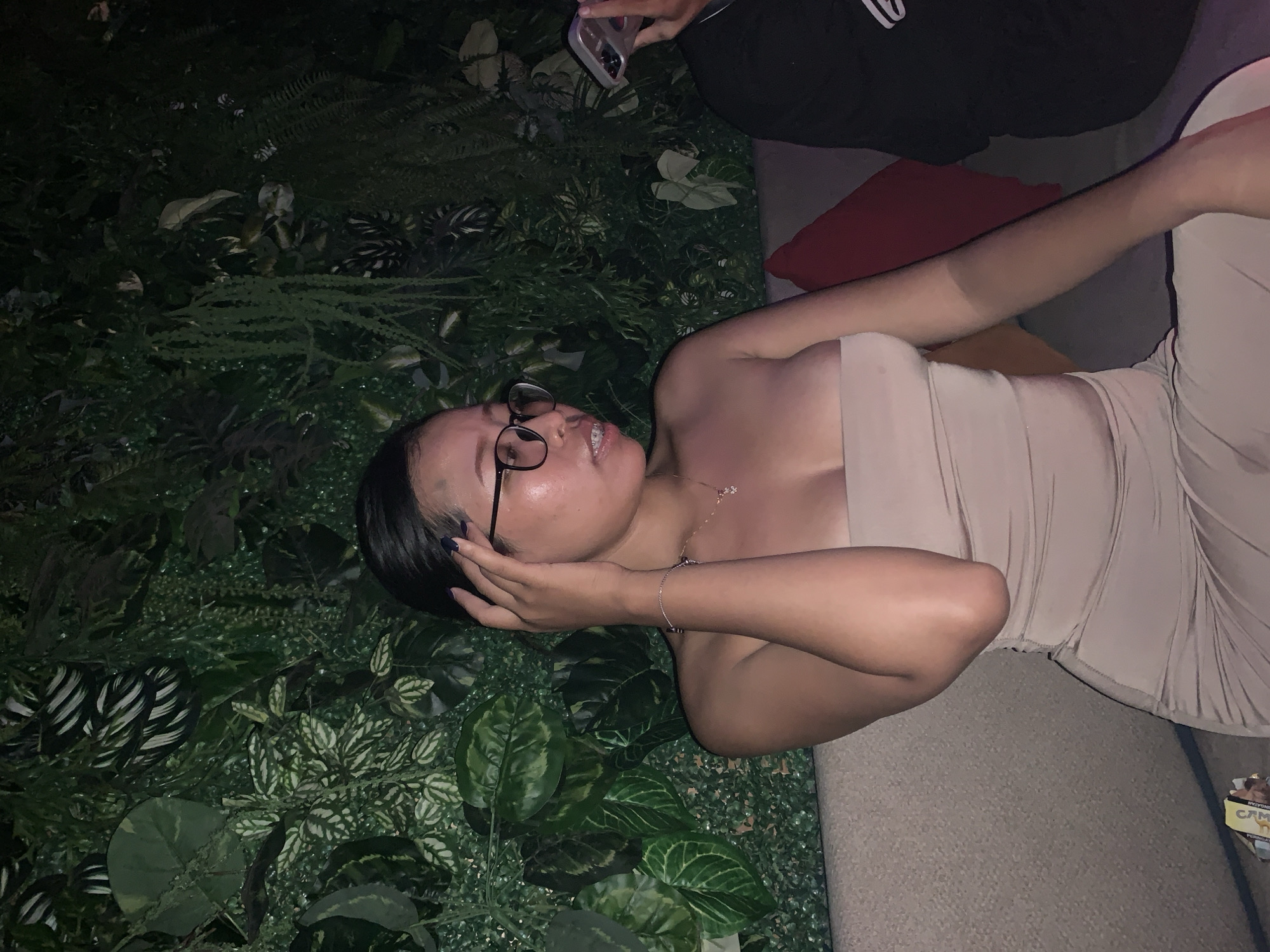 A young woman with dark hair and glasses is lying on a grey surface or couch, partly surrounded by lush green foliage. She is wearing a beige strapless dress. Her left hand is near her face. Above her, a person is holding a phone, presumably taking a picture. The setting appears to be outdoors or in a space decorated to mimic an outdoor environment. The location may be in North Kuta, Indonesia, based on additional information not visible in the image.
