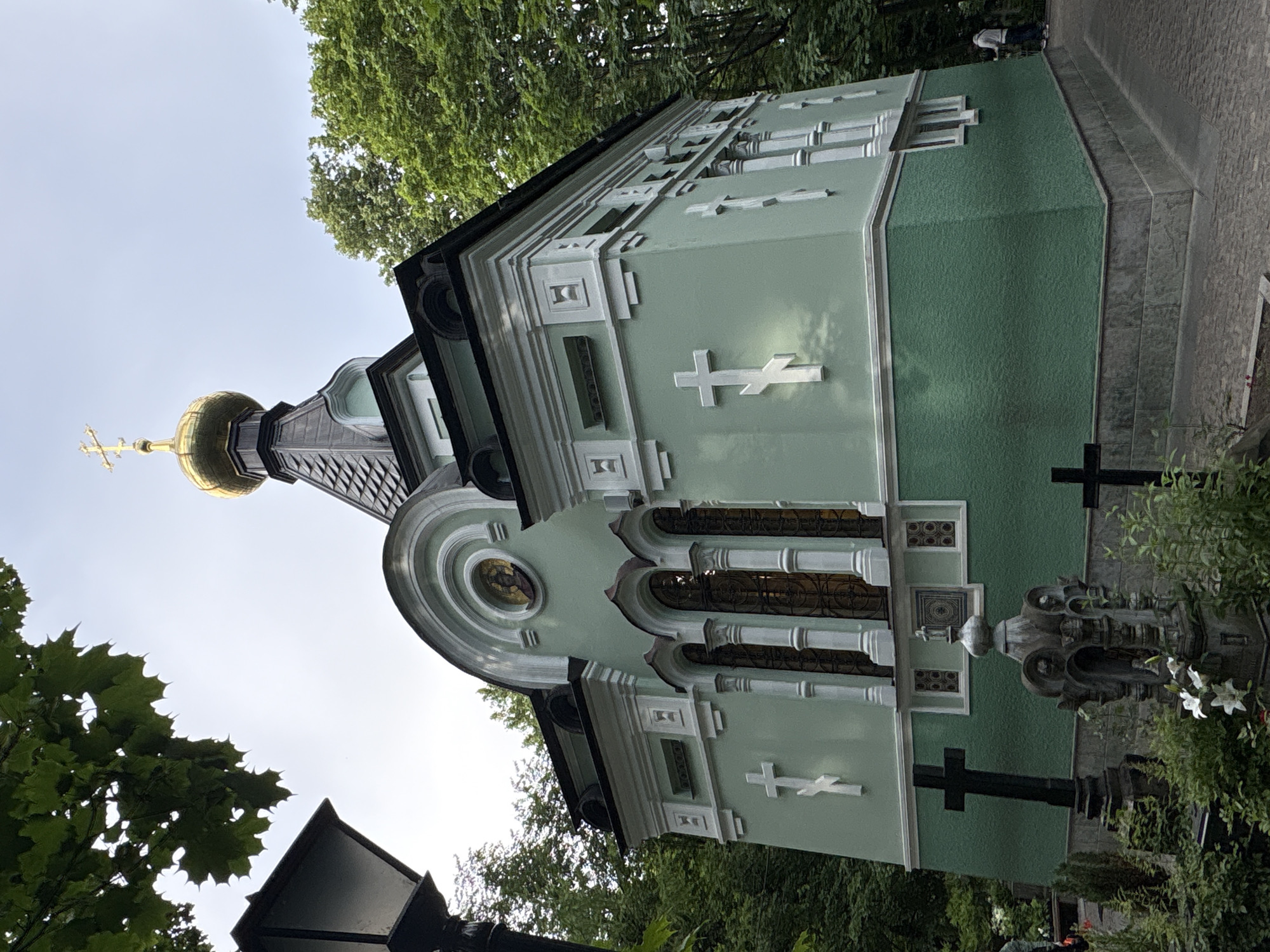 The image shows an exterior view of a small, green Orthodox chapel with a golden dome topped with a cross, situated in a cemetery. The chapel has white architectural detailing, including decorative molding and crosses mounted on its exterior walls. Several gravestones and funerary monuments are visible in the foreground, surrounded by lush greenery. In the background, mature trees frame the scene, and a lamppost stands to the left. The sky is overcast. The scene is located at Nikolskoe Cemetery, located within the Alexander Nevsky Lavra in Saint Petersburg, Russia. Two individuals are partially visible in the distance on the right, one walking away from the camera and the other stationary.