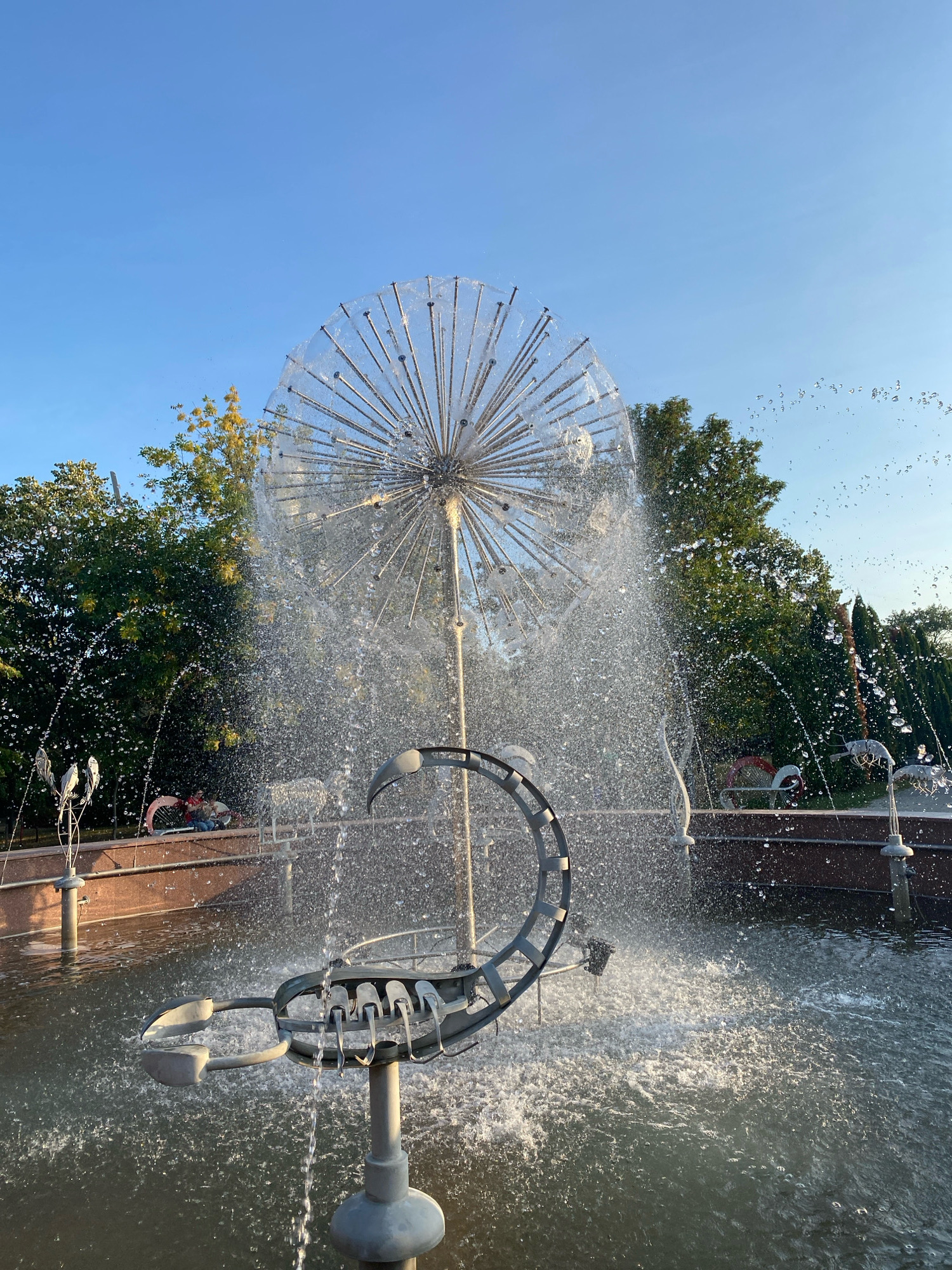 The image depicts a public fountain in Chișinău, Moldova, likely within a park or public square. The fountain features a large, spherical structure made of numerous metal rods, resembling a dandelion seed head, from which water cascades down. In the foreground, a metallic sculpture shaped like a scorpion's tail with claws is partially submerged in the fountain's pool. Water is actively spraying from various points of the fountain, creating a dynamic and wet environment. The background shows lush green trees and a clear blue sky. The ground around the fountain is paved with reddish-brown stone. The lighting suggests it is daytime, with the sun casting shadows. No people are clearly visible interacting with the fountain, though there might be distant figures or objects suggesting the presence of other park-goers or decorative elements within the pool. The overall scene is one of a well-maintained public water feature.