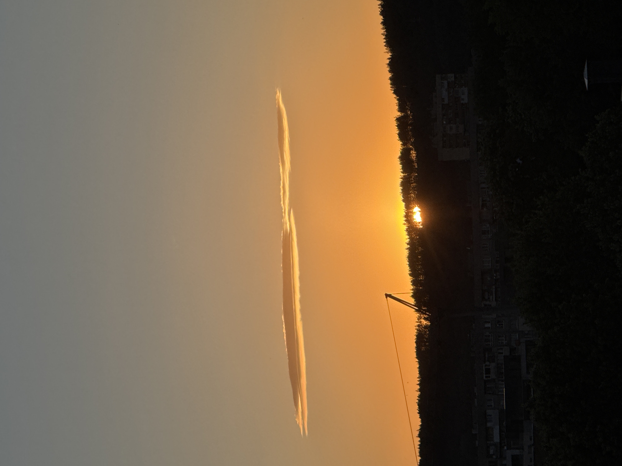 The image captures a sunset over the city of Pernik, Bulgaria, as viewed from a distance. The sky is a gradient of warm orange and soft gray, with a distinct, elongated cloud formation stretched horizontally across the middle of the frame. Below the sky, a dark silhouette of a tree-covered hill forms the backdrop. Emerging from the trees on the left is the structure of a construction crane, its arm angled upwards. The sun itself is partially obscured by the trees and the hill, appearing as a bright, luminous orb peeking through the foliage, casting a golden glow. In the lower half of the image, the dark outlines of buildings, likely apartment blocks, are visible amidst the dense foliage of trees. The scene is devoid of any visible people or direct interactions. The overall impression is one of a serene, urban twilight.