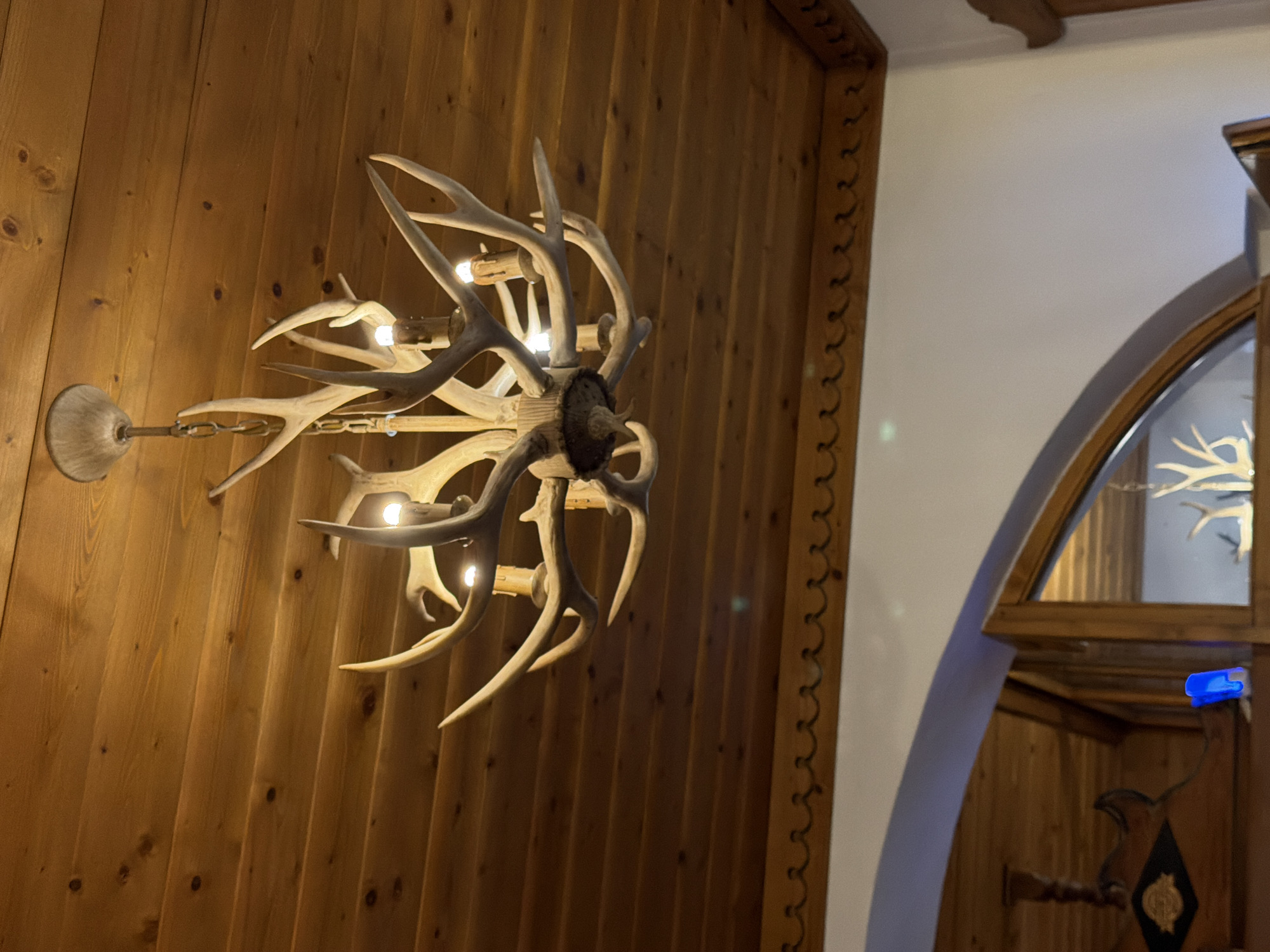 A chandelier made of deer antlers hangs from a wooden ceiling in Sauris di Sopra, Italy. The chandelier has multiple arms, each tipped with a light bulb designed to resemble a candle. The ceiling is made of horizontal wooden planks. Below the ceiling, a decorative wooden trim with a wavy pattern runs along the top of a white wall. An arched opening is visible, framed by wood, revealing a reflection of more antlers and a blue object inside.