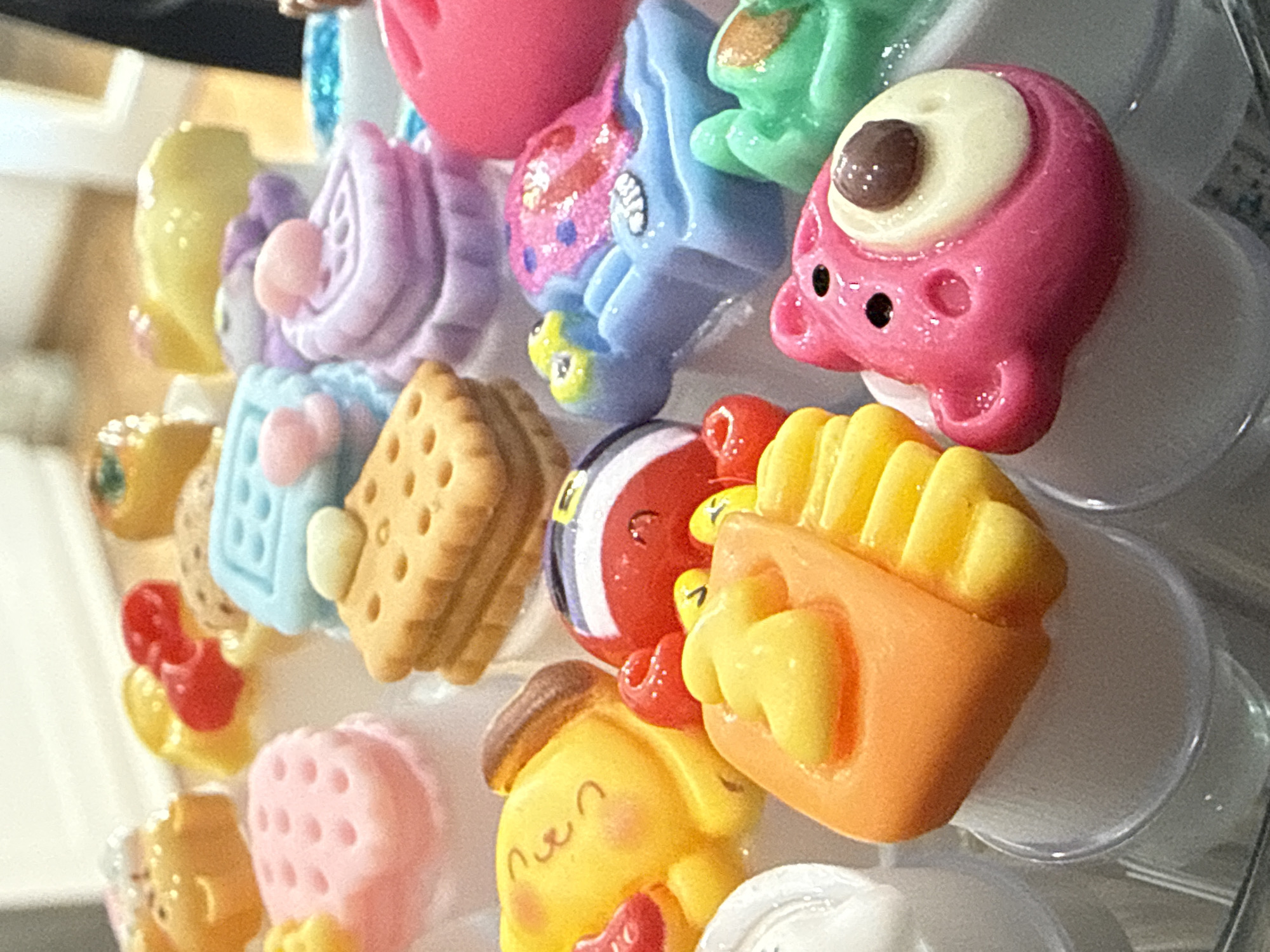 The image presents a close-up view of numerous small, colorful, cartoon-like figurines or charms, tightly arranged together on a display. These items, likely made of plastic or resin, exhibit a mix of vibrant and pastel colors such as pink, light blue, yellow, green, red, purple, and beige.
Prominent subjects include a pink, one-eyed creature with small black eyes, resembling an 'Among Us' character; a light blue cupcake decorated with pink hearts; a beige cracker or biscuit; and a light purple swirled object. Also visible are a light blue figure with glittery pink details and a yellow base, a green frog-like character, and a red and white striped ball. An orange container holds yellow french fries, and a yellow character with a brown beret and a smiling face, reminiscent of Pompompurin, is featured. Other items include a red bow, a pink square with perforated holes, and various other miniature shapes.
Many of the figurines are attached to clear plastic caps or bases, suggesting they are designed as collectible charms, pins, or decorative buttons. The objects are displayed statically, with no specific activity taking place. The background is softly blurred, indicating an indoor setting with ambient lighting. There is no visible text on any of the items. The overall impression is one of a playful and whimsical collection of novelty items. The location context for this scene is Chaikovsky, Russia.