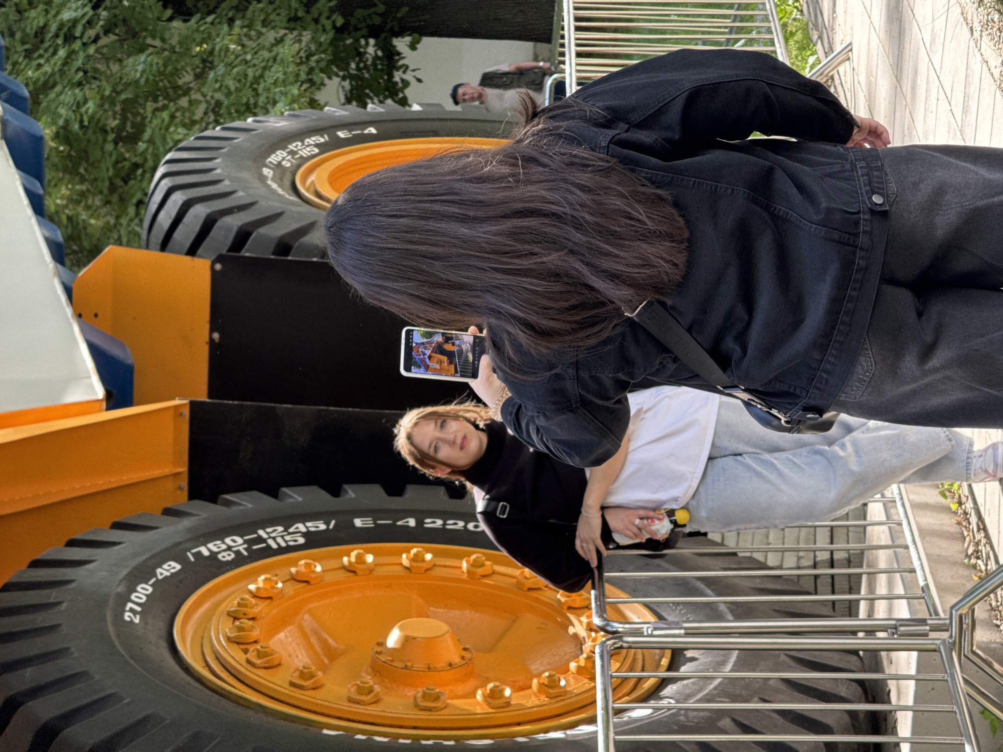 A young woman with long, dark hair, wearing a black denim jacket and dark grey pants, photographs a massive yellow and orange industrial vehicle with a smartphone. The vehicle's enormous tire, with the inscription "2700-49 / 1760-1245/ 8T-115 E-4220", is prominently featured. Behind the woman photographing, another young woman with light brown hair, wearing a black turtleneck and light blue jeans, leans against a metal barrier, looking towards the camera. She holds a small yellow object in her right hand. In the background, a man in a cap and vest stands near a tree. The scene appears to be outdoors, possibly in a public space or exhibition, with a metal fence in the foreground and trees in the background. The setting is consistent with the VDNKh (Exhibition of Achievements of National Economy) complex in Moscow, Russia, which often features large industrial exhibits.
