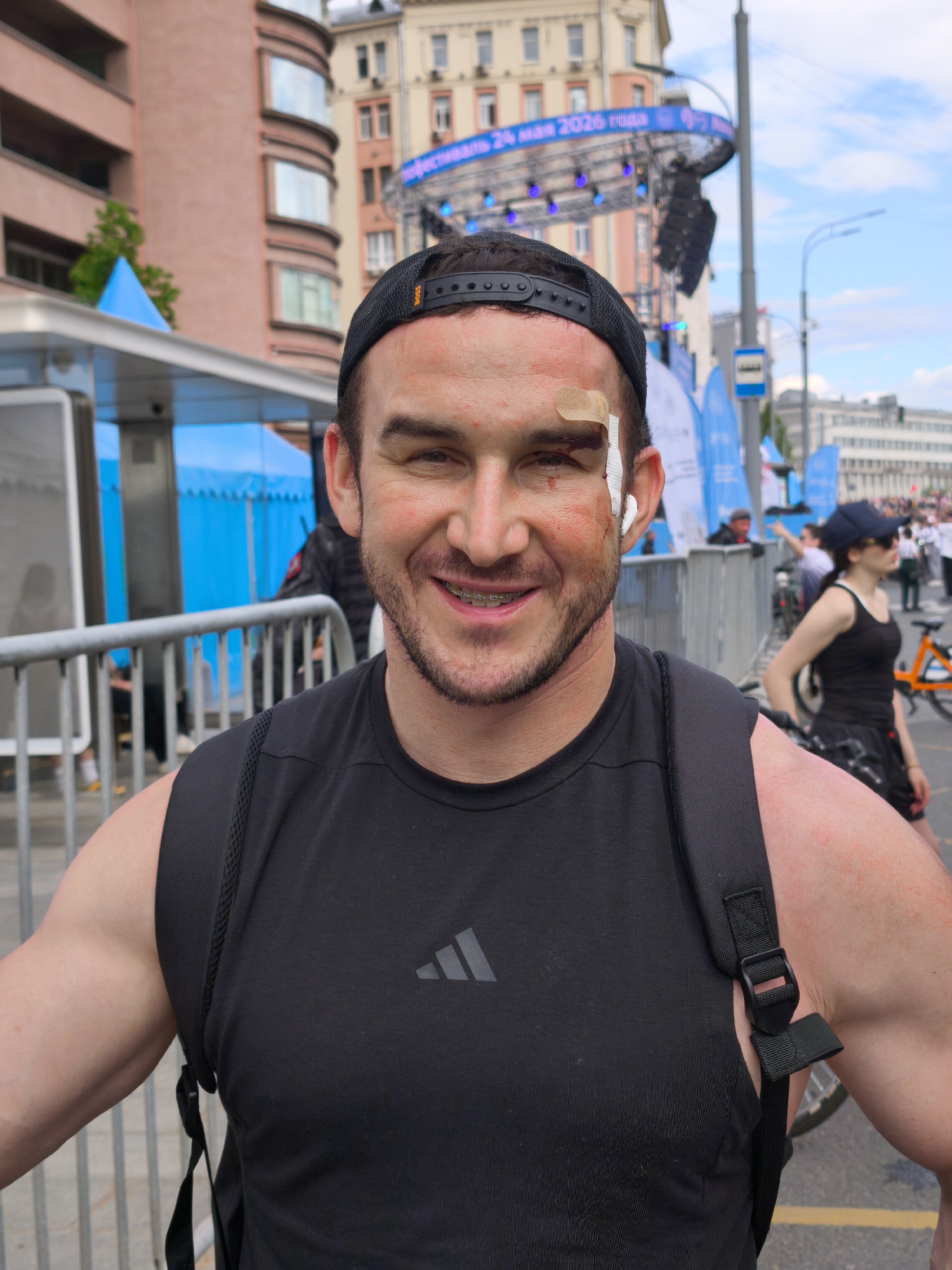 A medium shot captures a man smiling at the camera, wearing a black Adidas tank top and a black cap with a visible backpack. He has a bandage on his forehead and visible redness around his eyes and on his arms. He also has braces.
Behind him, there's a metal barricade and a bustling urban scene. A woman with a cap and sunglasses is visible in the background, along with a bicycle. Buildings line the background, and there's a stage with banners indicating an event, likely in Moscow, Russia, as evidenced by the Russian text. A bus stop sign is also visible.