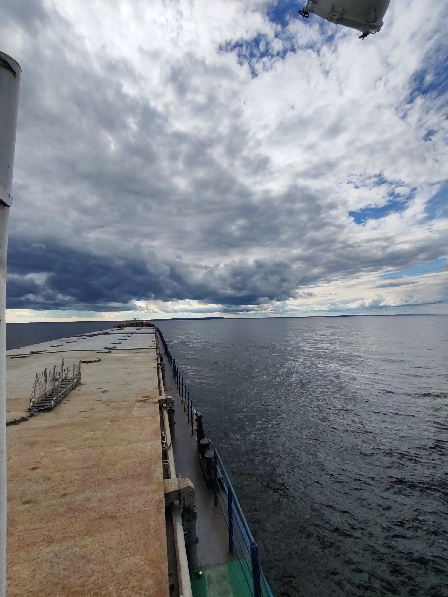 A wide-angle shot captures a view from a ship or pier, looking out towards a vast body of water under a cloudy sky. The foreground features a concrete surface with a metal walkway or railing along the right edge. In the distance, the horizon is a clear line separating the water from the sky, with landmasses faintly visible on either side. The sky is filled with a mix of dark, stormy clouds and brighter, scattered clouds. The water appears choppy with some ripples.