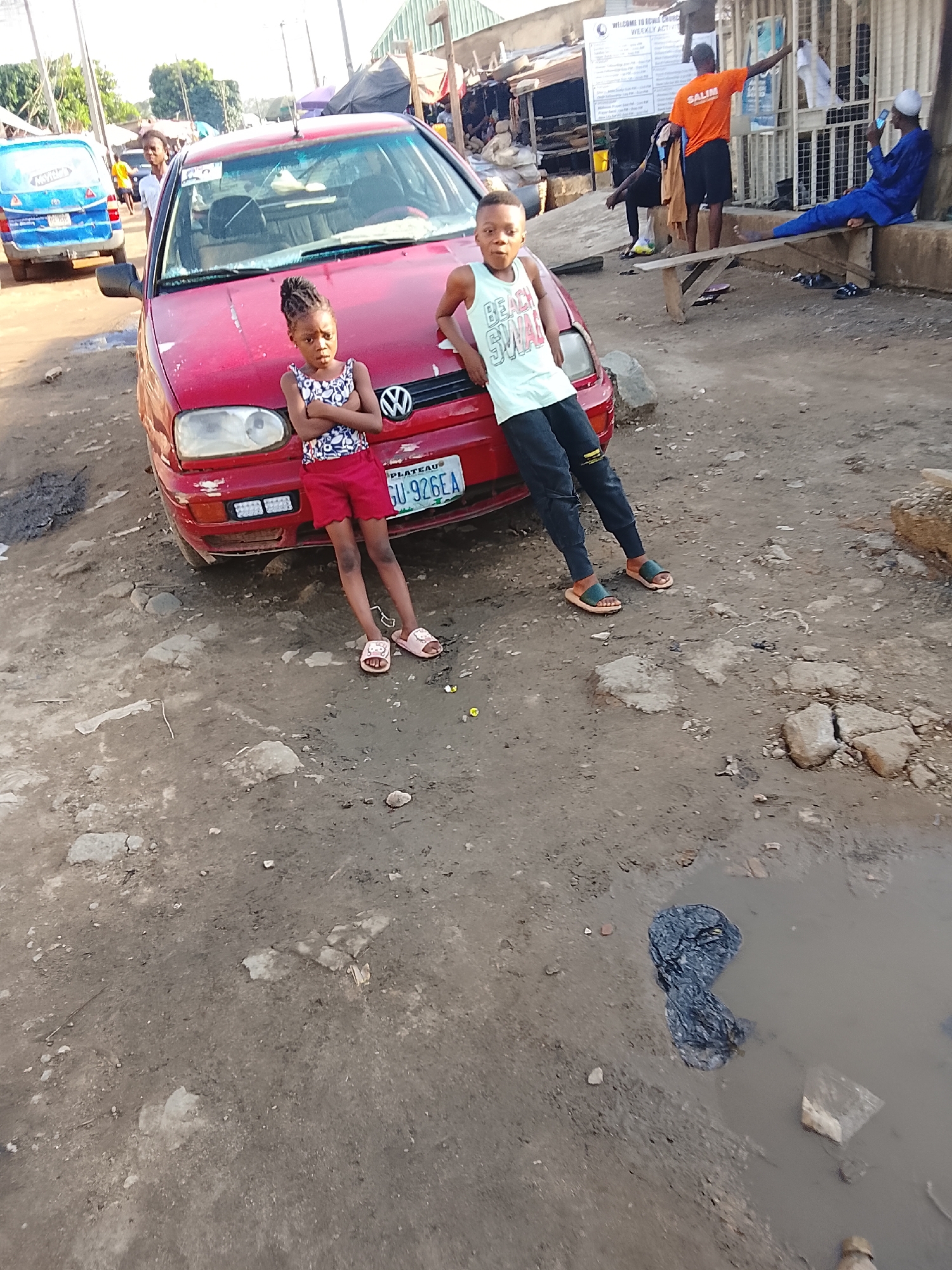 A low-angle shot depicts a dusty, somewhat dilapidated urban scene. Two children stand next to a red Volkswagen car, likely a Golf model, which appears to be older with some damage. The girl on the left wears a patterned top and red shorts, her arms crossed. The boy to the right has on a tank top and dark pants and is leaning against the car. The car's license plate reads "PLATEAU GU-926EA."
The surroundings are unpaved and rough, with dirt, stones, and a puddle containing dark debris visible in the foreground. In the background are structures and other people; some are seated on a makeshift bench, others walking around. There's a blue van parked in the distance to the left. A sign is visible on one of the buildings.
Based on the license plate and the general surroundings, this scene is likely from an urban area in Nigeria, possibly in or around the city of Abuja or Plateau State. The environment suggests a lower-income area.