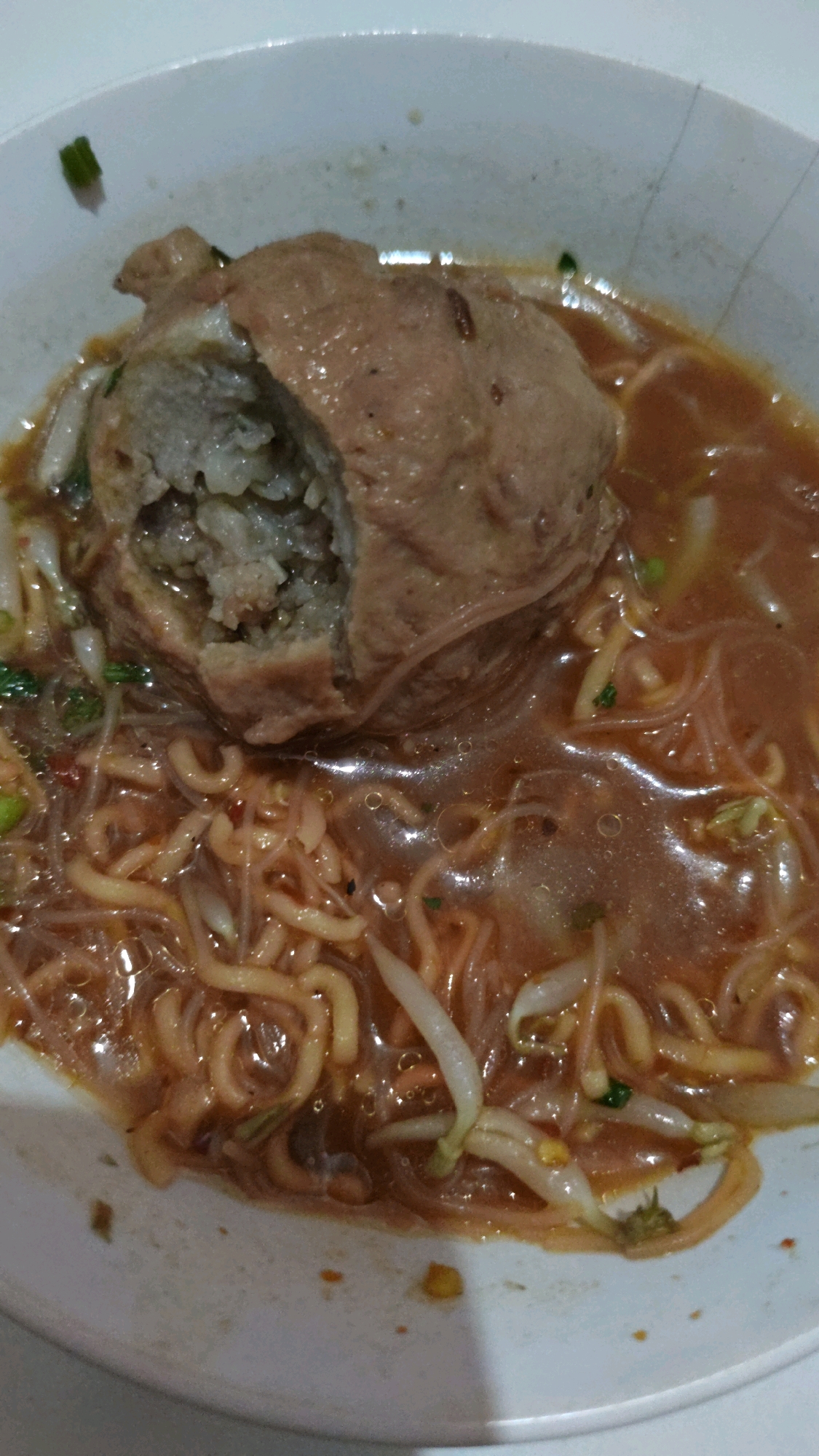 A close-up overhead shot shows a bowl of bakso, a popular Indonesian meatball soup. The dish features a large, spherical meatball, partially cut open to reveal its minced meat interior, resting in a rich, reddish-brown broth. The broth is filled with thin, yellow noodles and white bean sprouts, with small flecks of green garnish scattered throughout. The bowl is white ceramic, with its rim visible at the top and bottom of the frame. The lighting is direct, creating some glare on the glossy surface of the broth. The background is indistinct and appears to be a light-colored, textured surface, possibly a table. This dish is commonly found in street food stalls and restaurants throughout Cimahi, Indonesia, and is a staple of Indonesian cuisine.