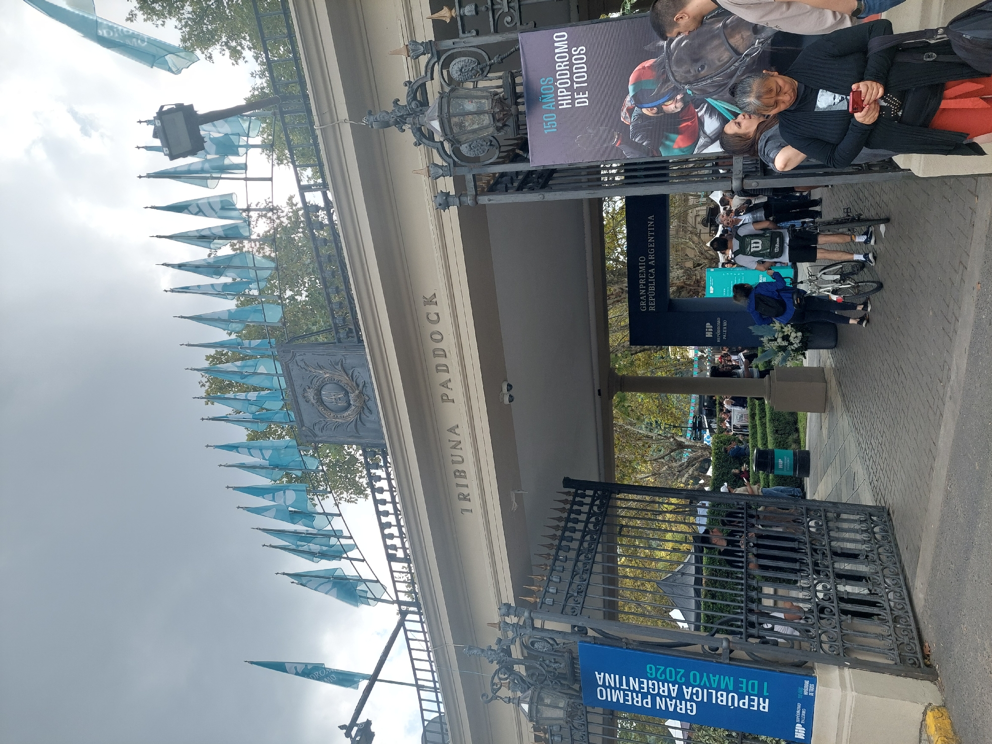 The image captures an outdoor scene at the ornate entrance to what appears to be a grandstand or paddock area of a racecourse, under an overcast sky. A large, dark, intricately designed wrought-iron gate, partially open, dominates the lower part of the frame, leading to a paved walkway.
The main building, identified by the inscription "TRIBUNA PADDOCK" on its facade, features a large arched entryway and classical architectural elements. Numerous light blue flags or pennants, some bearing partial text like "DROM MO" (likely part of "HIPODROMO"), are strung across the entrance structure, billowing slightly in the breeze.
Several people are visible in various parts of the frame. In the mid-ground, a woman in a black shirt and dark pants is seated, looking at a red smartphone. She is near a large vertical banner on the right, which advertises "150 AÑOS HIPÓDROMO DE TODOS" (150 Years Hippodrome for Everyone) and displays an image of a jockey on a horse. Further back, other individuals are gathered or walking, some with bicycles, indicating a public event or gathering.
Another prominent vertical banner on the left side of the entrance announces "GRAN PREMIO REPÚBLICA ARGENTINA 1 DE MAYO 2026" (Grand Prize Republic of Argentina May 1, 2026), also displaying "HIPODROMO PALERMO" and "HIP" branding. This clearly places the event at the Hipódromo de Palermo in Buenos Aires, Argentina. A "Wilson" logo is visible on a backpack worn by one person. The overall atmosphere suggests an active event day at the racetrack.
