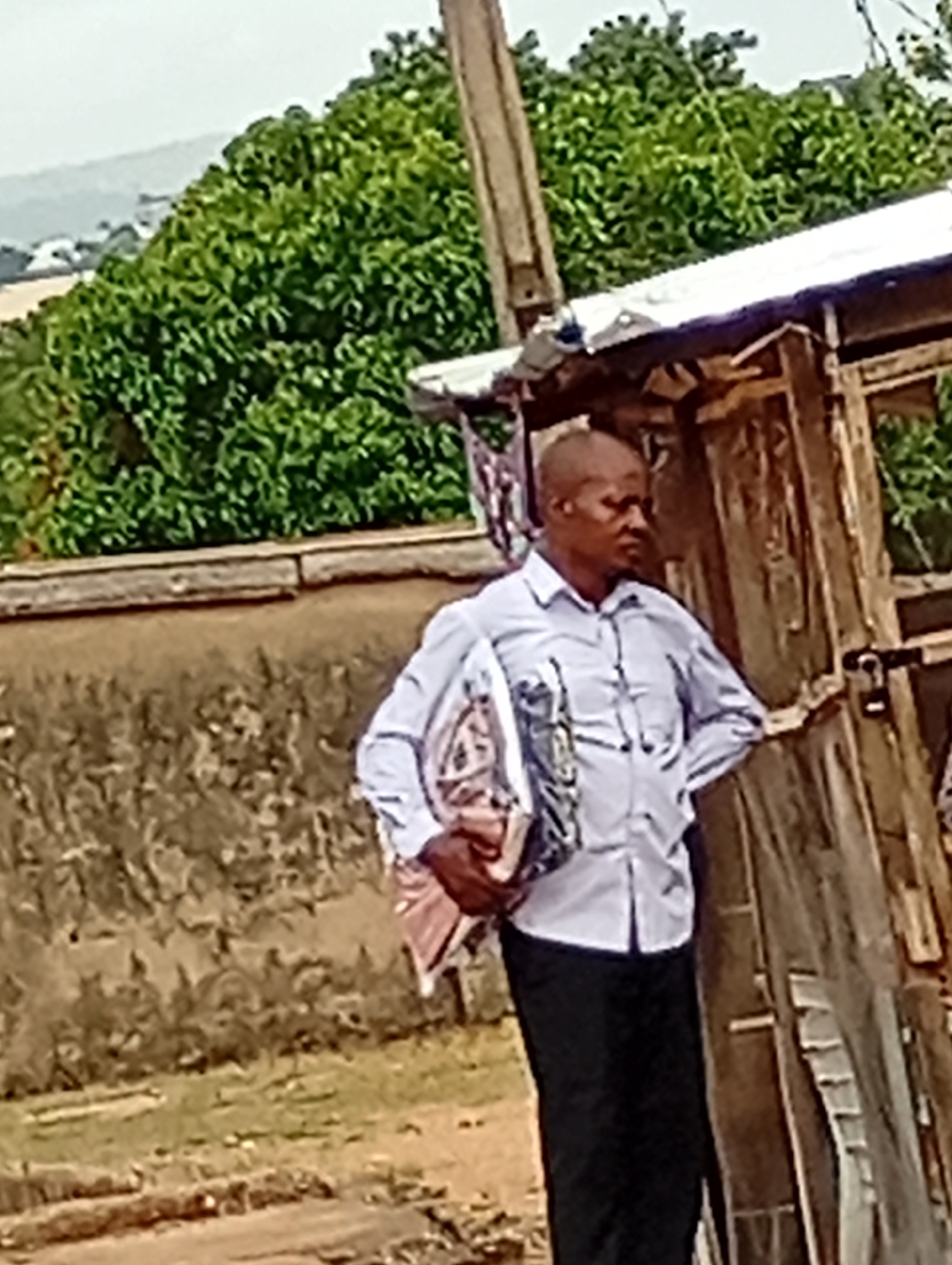 A man stands on a street in Adankolo, Nigeria, holding a package. He wears a light blue button-down shirt and dark trousers. He has a shaved head. Behind him is a weathered wooden structure, possibly a shed or shop. A concrete wall runs along the left side of the frame, and lush green trees fill the background. In the distant background, a hazy horizon is visible.