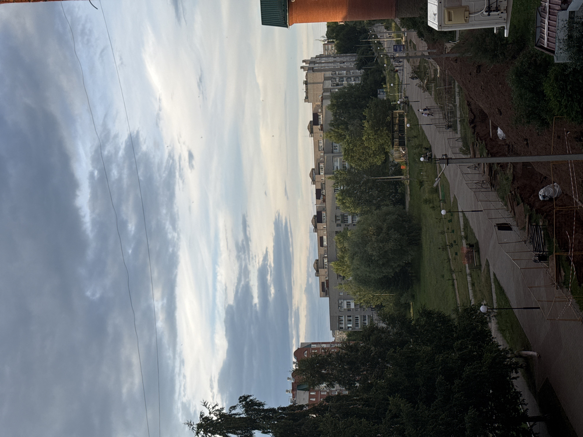 This media file captures an outdoor scene in Chaikovsky, Russia, likely during the late afternoon or early evening. The perspective is from a high vantage point, looking down and across a residential area.
The primary subjects are several multi-story apartment buildings, typical of urban or suburban environments in Russia. The buildings vary in architectural style and color, with some featuring brick facades and others painted in lighter tones. They are nestled amongst mature trees, which are in full leaf, suggesting a warm season like summer or early autumn.
A paved walkway or road runs through the scene, bordered by green spaces and more trees. Several lampposts are visible along the walkway, indicating it is a public area, possibly a park or pedestrian zone within the residential complex. There are a few people walking along the path, their presence adding a sense of everyday life to the scene.
Construction or repair work is evident in some areas, with piles of dirt and temporary fencing visible. This suggests ongoing urban development or maintenance.
The sky is a prominent element, filled with dramatic clouds that are a mix of dark grays and bright whites, indicating a dynamic weather system. The lighting suggests the sun is low in the sky, casting long shadows and creating a somewhat moody atmosphere.
There is no visible text in the image. The overall impression is a snapshot of daily life in a Russian city, with a blend of residential buildings, natural elements, and signs of human activity and infrastructure.