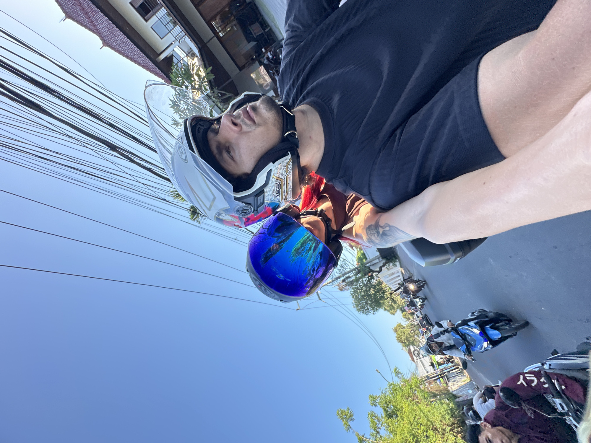 A medium shot depicts a man riding a motorcycle through the streets of North Kuta, Indonesia. He wears a black t-shirt and is equipped with two motorcycle helmets: a white helmet with a clear visor, and a blue reflective helmet. The image is taken from a low angle, showing a clear blue sky above and numerous power lines crisscrossing overhead. The surrounding environment includes buildings and lush greenery, indicative of an urban area. Other motorcyclists and vehicles are visible on the road, suggesting a typical urban scene.