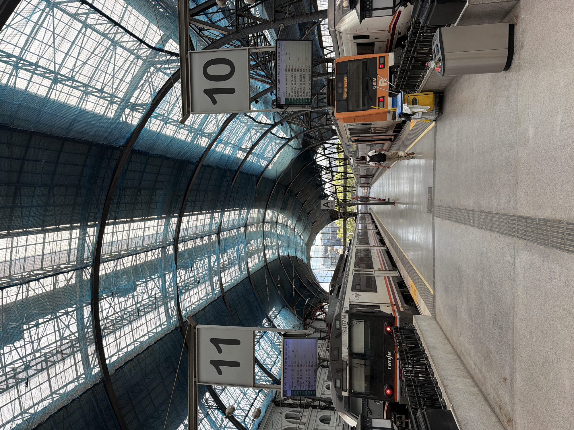 This is a photograph taken inside a train station with a high, arched glass roof supported by dark metal beams. The station appears to be a major hub, with multiple tracks and platforms. Two trains are visible at the platforms. The train on the left is black and white with "renfe" branding. The train on the right is orange and white, also with "renfe" branding. Both trains are stopped at the platform.
There are digital departure boards mounted on poles above each platform. The board above platform 11 shows "11" prominently, and a digital display listing departure times and destinations, including "VILANOVA I LA GELTRU". The board above platform 10 shows "10" prominently, and a similar departure board.
A lone individual, a person wearing a light-colored shirt, khaki pants, and a backpack, is walking away from the camera down the platform between the two trains. A yellow cleaning cart with mops and cleaning supplies is parked next to the orange train. A silver trash receptacle is situated on the platform to the right.
The environment is clean and well-maintained. The lighting is natural, filtering through the glass roof, creating a bright and airy atmosphere. The architecture of the station is grand and industrial, suggesting a historic and important transportation facility. The image is likely taken at Barcelona Sants Station, a major railway hub in Barcelona, Spain, known for its distinctive arched glass roof.