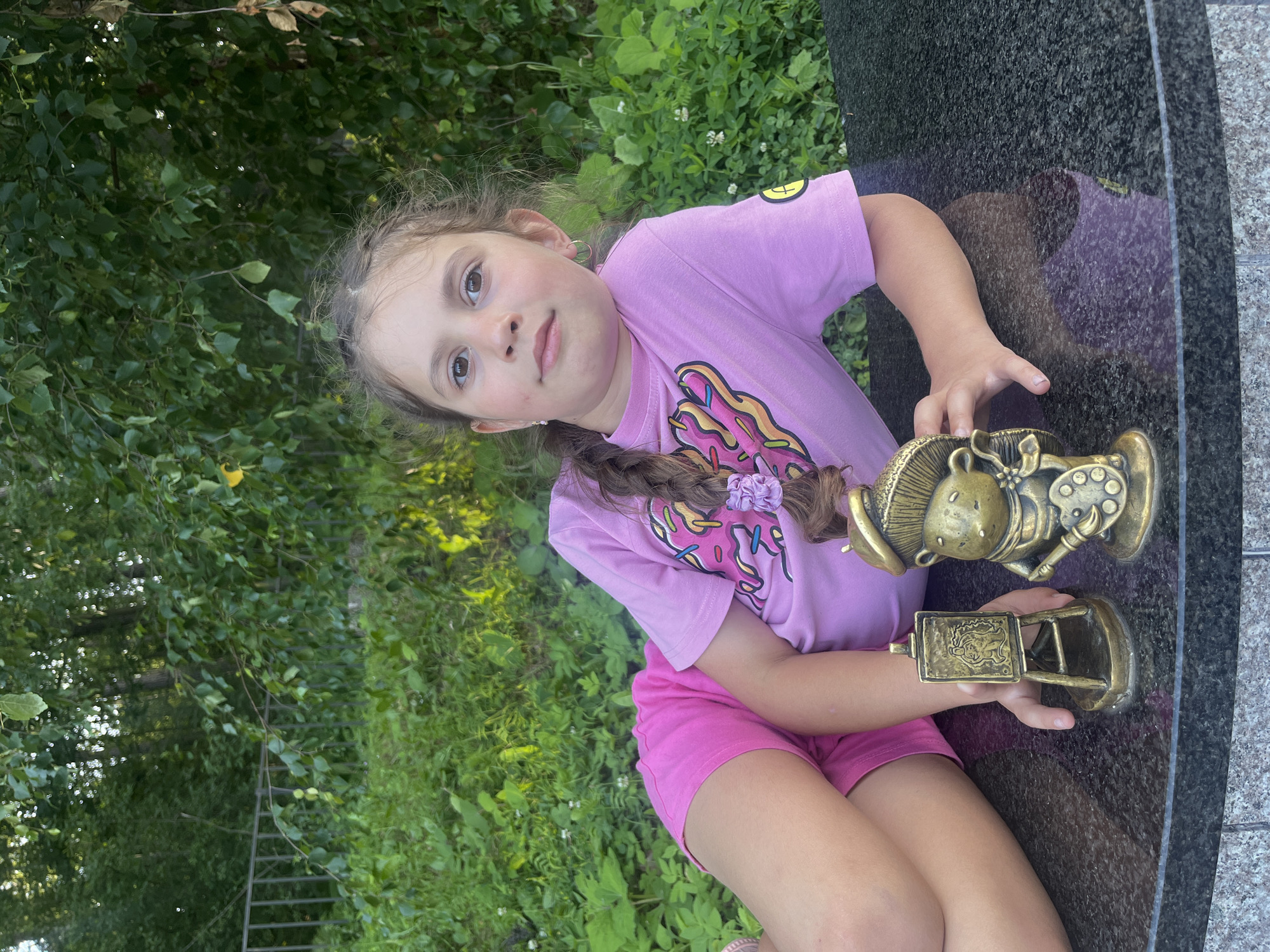 In this outdoor media file, a young girl is the central subject. She is sitting on a dark, polished surface, likely a monument or a stone bench, with lush green foliage and trees in the background. The girl is wearing a bright pink t-shirt with a graphic design, and pink shorts. Her hair is braided, and she has a scrunchie in her hair. She is holding two golden-colored objects, which appear to be small statues or trophies. One is a figurine of a character, possibly a mouse, dressed as an artist, holding a palette and brush. The other is a smaller rectangular item that resembles a mini easel or a plaque with an embossed image.
The lighting suggests it's daytime, possibly late afternoon or early evening due to the soft light and the presence of shadows. The overall mood seems relaxed and possibly celebratory, given the presence of the trophies. The background with trees and greenery indicates a natural or park-like setting. The polished stone she's sitting on might be part of a memorial or a public art installation. There is no visible text in the image. The girl's expression is calm and her gaze is directed towards the viewer.