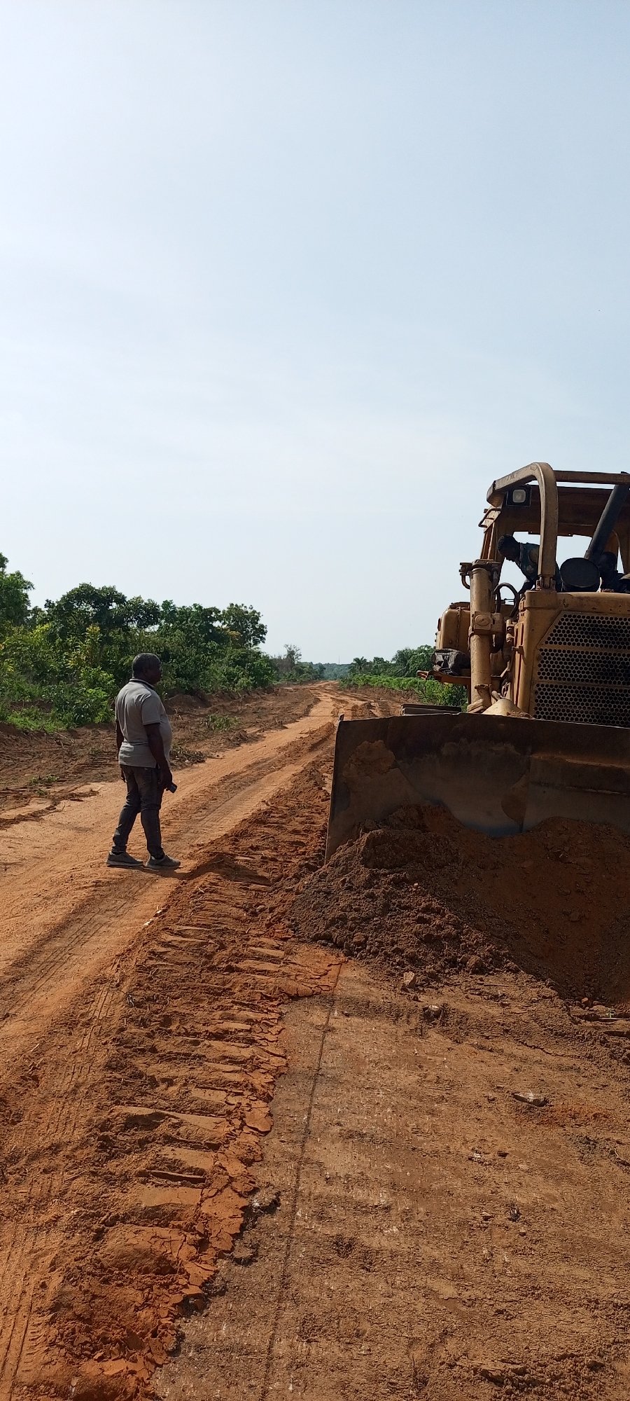 The image captures a scene of land clearing or road construction in a rural area of Otunade, Nigeria. A wide, unpaved road of reddish-brown earth, marked with tire and track impressions, extends from the foreground into the distance.
In the left mid-ground, a man stands on the dirt road, facing away from the viewer and looking towards the heavy machinery. He is wearing a grey short-sleeved shirt, dark pants, and sneakers, appearing to supervise or observe the work.
Dominating the right side of the frame is a large, yellow bulldozer, with its front blade lowered and pushing a substantial pile of freshly excavated soil. A person can be indistinctly seen inside the open cabin of the bulldozer, operating the machinery.
The background features abundant green foliage, consisting of trees and bushes, lining both sides of the developing road. The sky above is clear and light blue, indicating a bright, sunny day. The scene conveys an ongoing infrastructure project in a natural, undeveloped setting. No discernible text is visible in the image.