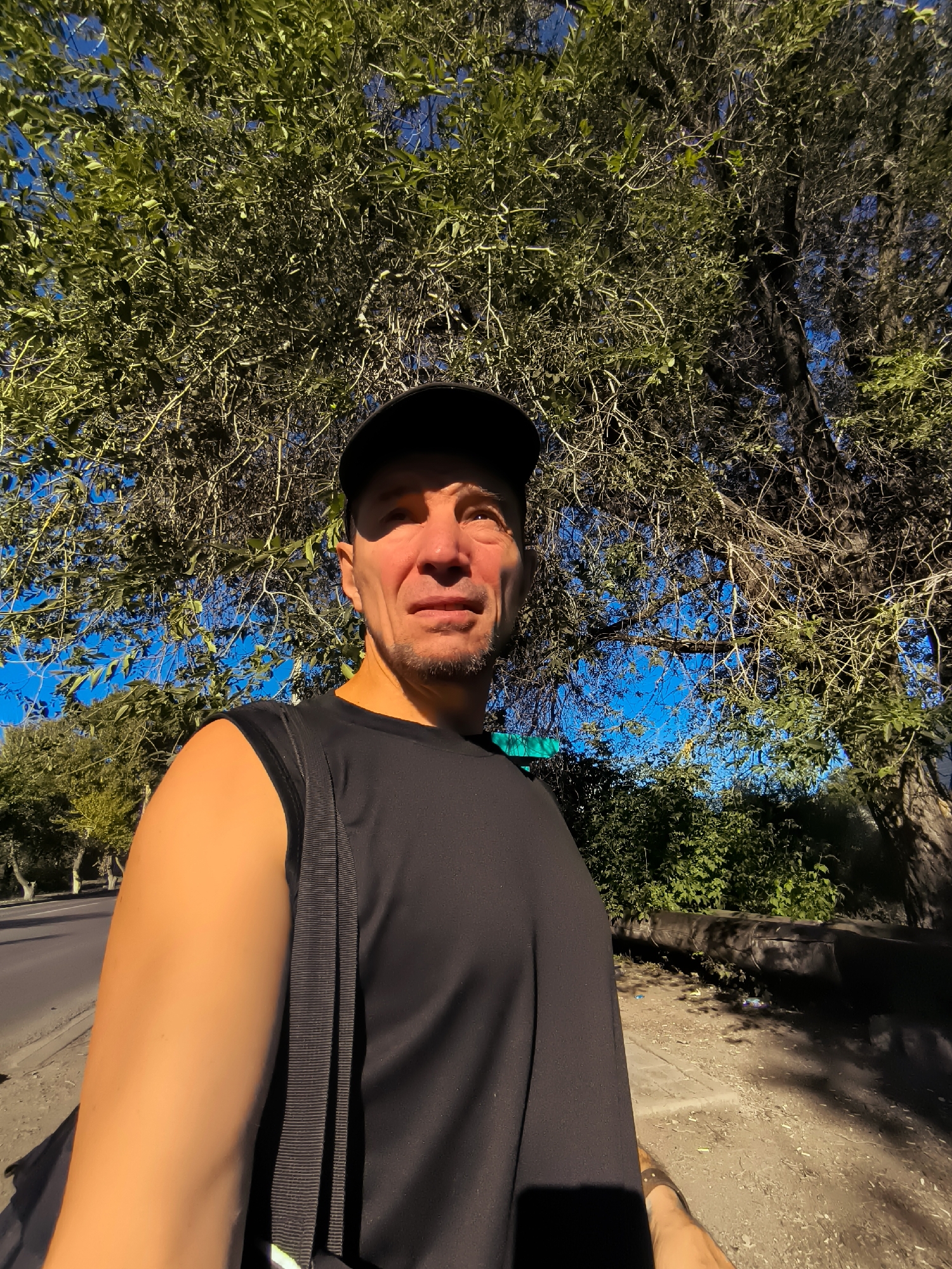 A man wearing a black baseball cap and a black sleeveless shirt is standing outdoors, with a tree and blue sky in the background. He is carrying a black bag over his shoulder. Sunlight casts shadows on the ground and highlights the man's face. To the left of the man, a paved road is visible with some trees lining the side. The scene appears to be in an urban or suburban area, possibly in or near Qarağandı city administration, Kazakhstan.