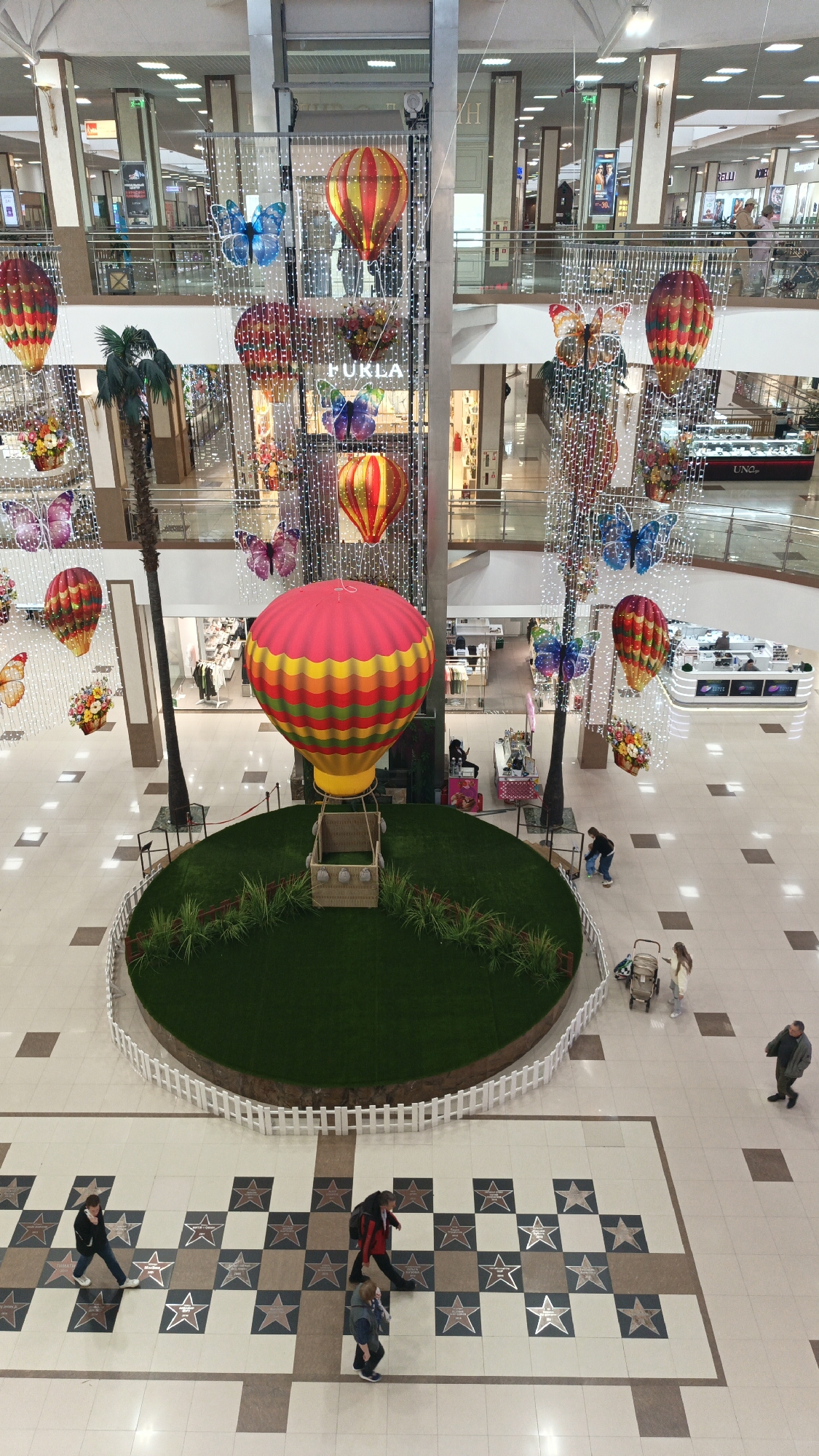The image depicts the interior of a multi-story shopping mall. Dominating the scene is a large hot air balloon decoration placed on a circular patch of artificial grass enclosed by a white picket fence. Surrounding the balloon are decorative butterflies, flower arrangements, and smaller hot air balloons suspended from the ceiling. A palm tree stands adjacent to the balloon display.
Several people are visible on the ground floor. One person is bending over near the balloon, while a woman and child with a stroller are standing nearby. A man walks toward the edge of the frame. A group of three people are walking over a tiled area with star-shaped inlays. Balconies overlook the scene on multiple upper levels. Retail stores are visible in the background, with one labeled "Furla." The general lighting is bright and the overall environment appears to be clean and well-maintained.
The location of the image is the Galeria Krasnodar shopping mall, in Krasnodar, Russia.