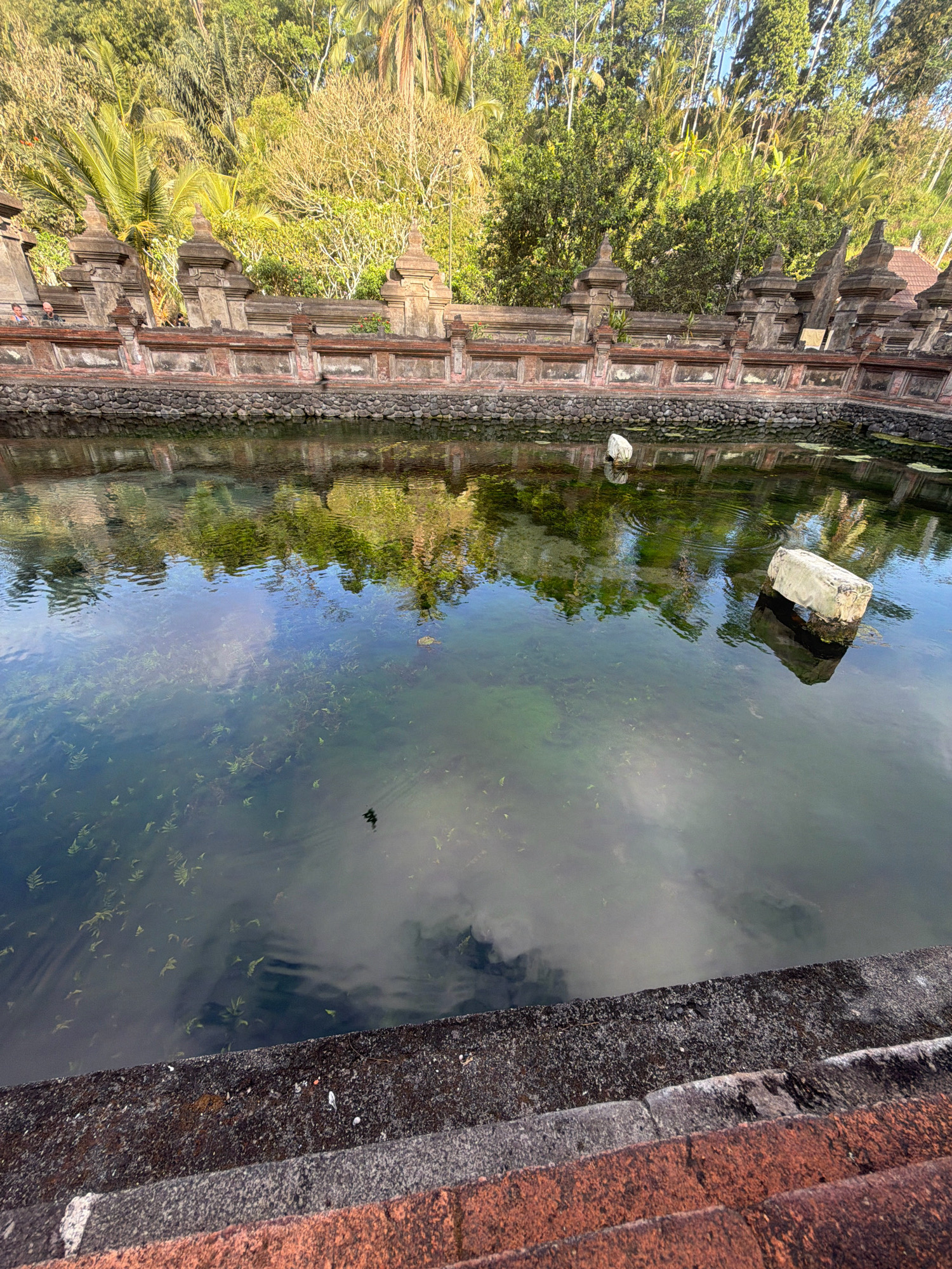 The image depicts a tranquil pool of water at the Tirta Empul Temple in Tampaksiring, Indonesia. The water is clear, revealing aquatic plants growing on the bottom. Reflections of the surrounding lush vegetation, including palm trees and other tropical foliage, are visible on the water's surface, along with the blue sky and white clouds.
The pool is bordered by a stone wall adorned with decorative architectural elements characteristic of Balinese temples. This wall is built with rough-hewn stones at its base and a more refined brick structure above, featuring segmented pilasters topped with finials.
Two large, light-colored rectangular objects, possibly weathered stones or ancient artifacts, are partially submerged in the water, creating ripples. The foreground shows the edge of the pool, constructed with rough-hewn reddish-brown stones and a darker, textured material. The overall scene conveys a sense of serenity and spiritual significance, typical of a sacred water temple. The lighting suggests it is either early morning or late afternoon, with the sun casting a warm glow on the landscape.