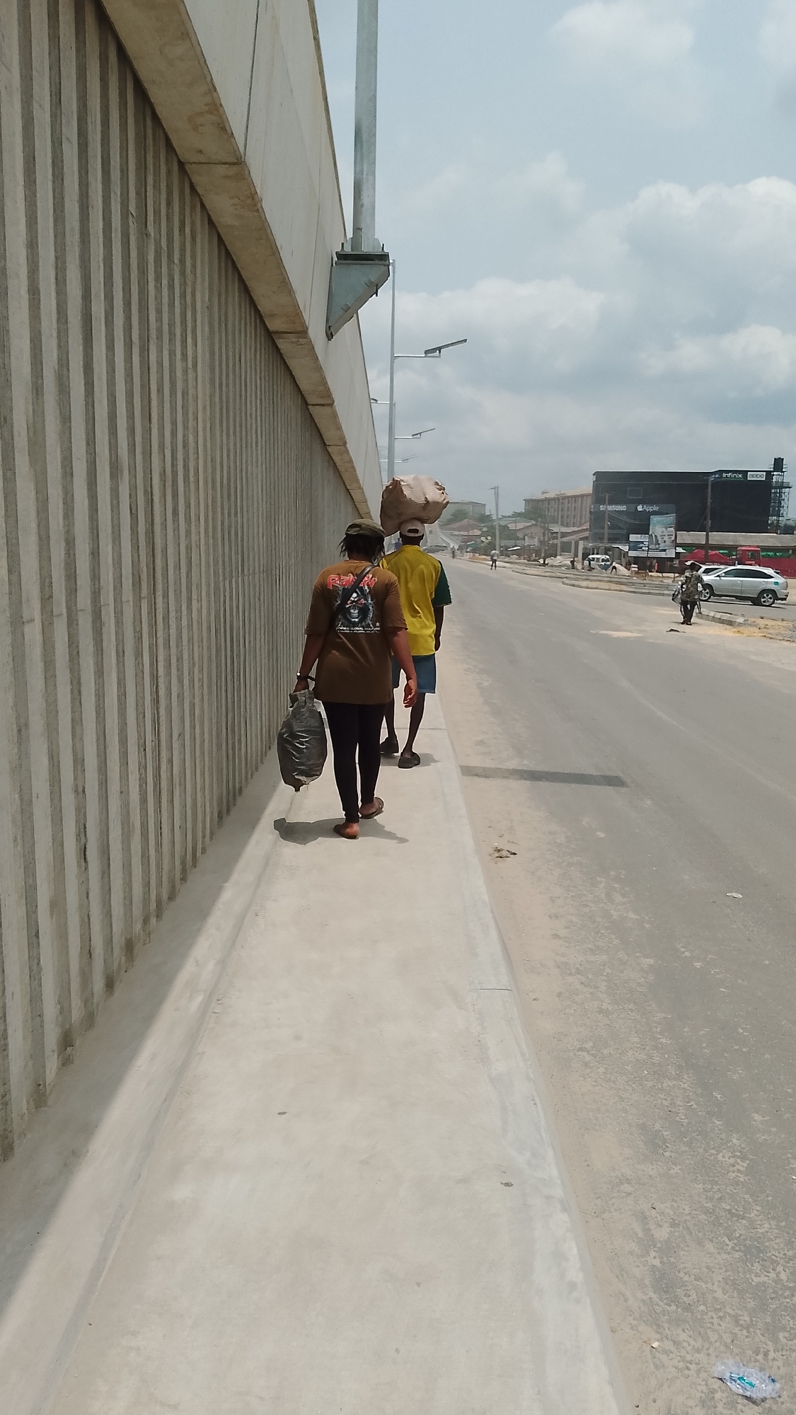The image shows two people walking along a concrete sidewalk in Effurun, Nigeria. A woman in a brown t-shirt and black leggings is carrying a black plastic bag. A man in a yellow t-shirt and blue shorts is carrying a large bag on his head. They are walking away from the camera, parallel to a tall concrete wall. Beyond them is a street with parked cars, pedestrians, and a building with a billboard advertising electronic devices. The sky is partly cloudy. Streetlights are visible along the roadside.