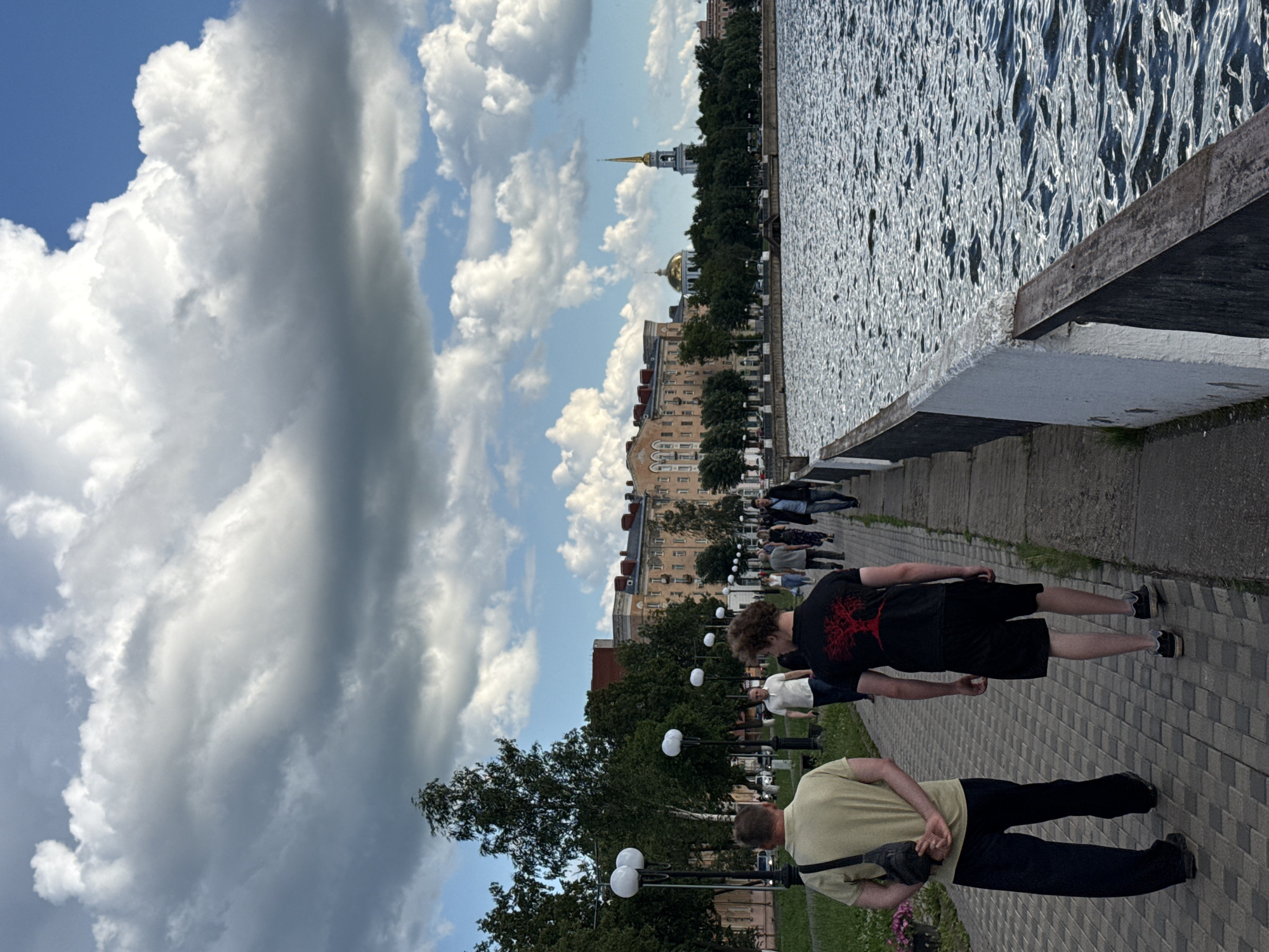 The image captures a scene along a waterfront promenade in Votkinsk, Russia. The sky is filled with dramatic, volumetric clouds against a clear blue sky, suggesting a dynamic weather pattern, possibly mid-afternoon. The sun intermittently breaks through, creating shimmering patterns on the rippling water.
The foreground features a paved walkway with a distinct diamond-patterned brickwork. Two young men are walking away from the camera along this path. The man closer to the foreground wears a light yellow t-shirt and dark trousers, with a black crossbody bag slung over his shoulder. The man in front of him is dressed in a black t-shirt with a red graphic and black shorts. Both appear to be casually strolling.
Further along the promenade, a line of people are walking in the same direction, creating a sense of a relaxed, public gathering. Beyond the promenade and a line of lush green trees, several multi-story buildings with traditional architecture line the embankment. Dominating the skyline in the distance are the golden dome of a church and a tall, slender bell tower with a spire, characteristic architectural elements of many Russian towns.
The overall mood is one of peaceful recreation, with people enjoying the waterfront and the scenic views. The interplay of light and shadow on the water and clouds adds a sense of depth and atmosphere to the scene. No visible text is present in the image.