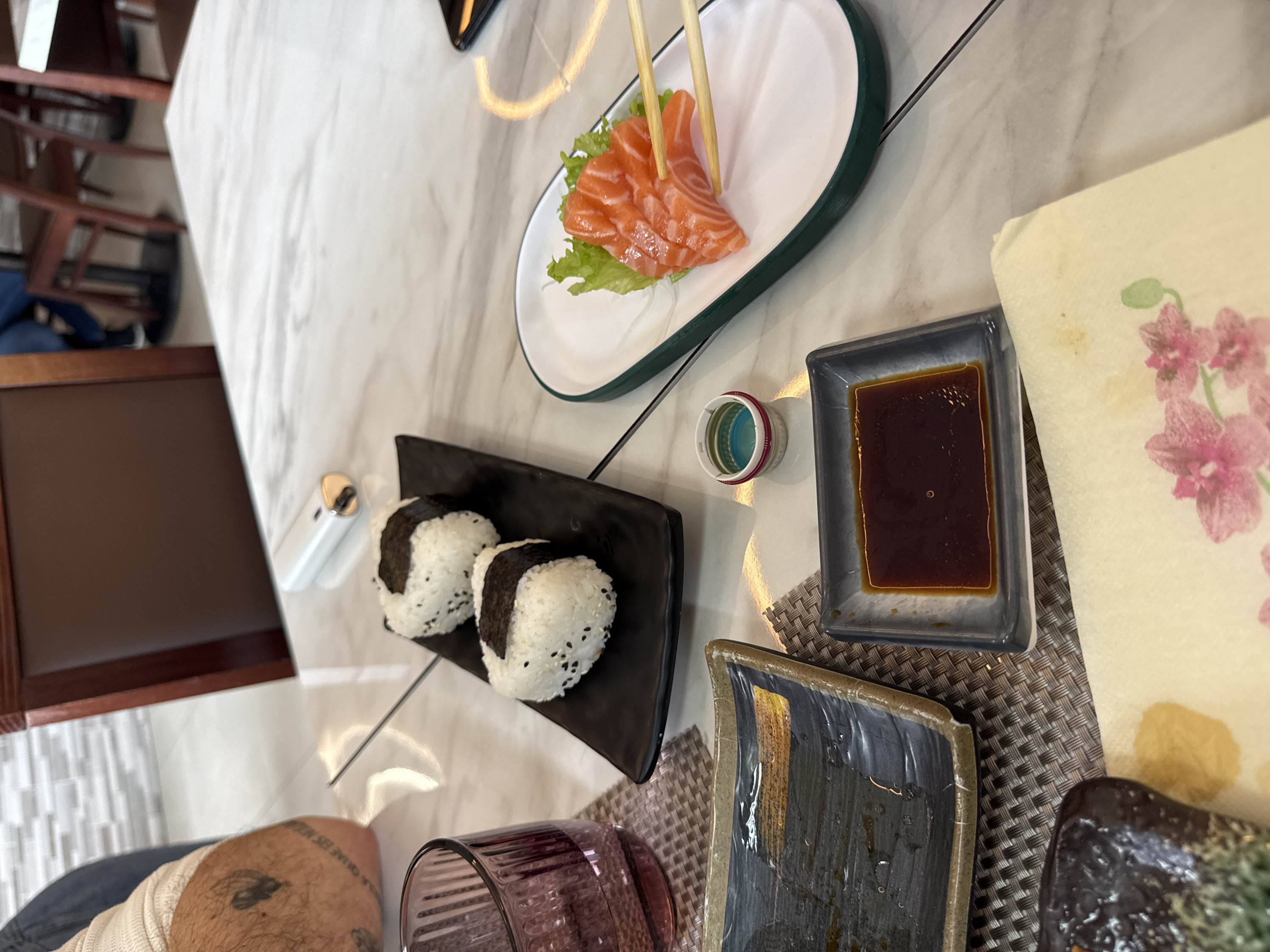 The image depicts a close-up, first-person view of a dining table in a restaurant in Milan, Italy. The scene shows various Japanese food items set on a light-colored, marble-patterned tabletop.
In the foreground on the left, a portion of a person's forearm, covered by a light-colored waffle-knit shirt, rests on the table. A dark tattoo is visible on the exposed part of the forearm. Below the arm, a ribbed, dark translucent glass sits on a brown woven placemat, partially filled with liquid. To the right of the glass, an empty, rectangular dark ceramic plate with a textured brown rim and a horizontal gold stripe is visible, showing residual moisture. In the immediate foreground to the right, a light cream-colored paper napkin with a pink floral design is placed.
Centrally positioned on the table are two main dishes. A black rectangular plate holds two triangular onigiri (rice balls) wrapped with dark nori (seaweed) and sprinkled with black sesame seeds. To the right of the onigiri, on a white oval plate with a dark green rim, several slices of orange salmon sashimi are arranged on a bed of green lettuce. A pair of light-colored wooden chopsticks is actively gripping one piece of salmon sashimi from this plate.
Other items on the table include a small, rectangular dark gray ceramic dish filled with dark brown liquid (likely soy sauce) below the onigiri plate. Beside this dish, a small, multi-colored plastic cap, appearing to contain a small amount of blue substance, is present. Behind the onigiri plate, a white and gold electronic vape device or lighter rests on the tabletop. The corner of a dark mobile phone is visible at the far right edge of the table.
The background shows blurred elements of the restaurant interior, including brown wooden chairs with upholstered backs and a light-colored tiled wall with a horizontal brick pattern. The floor appears to be a continuation of the light-colored tile.