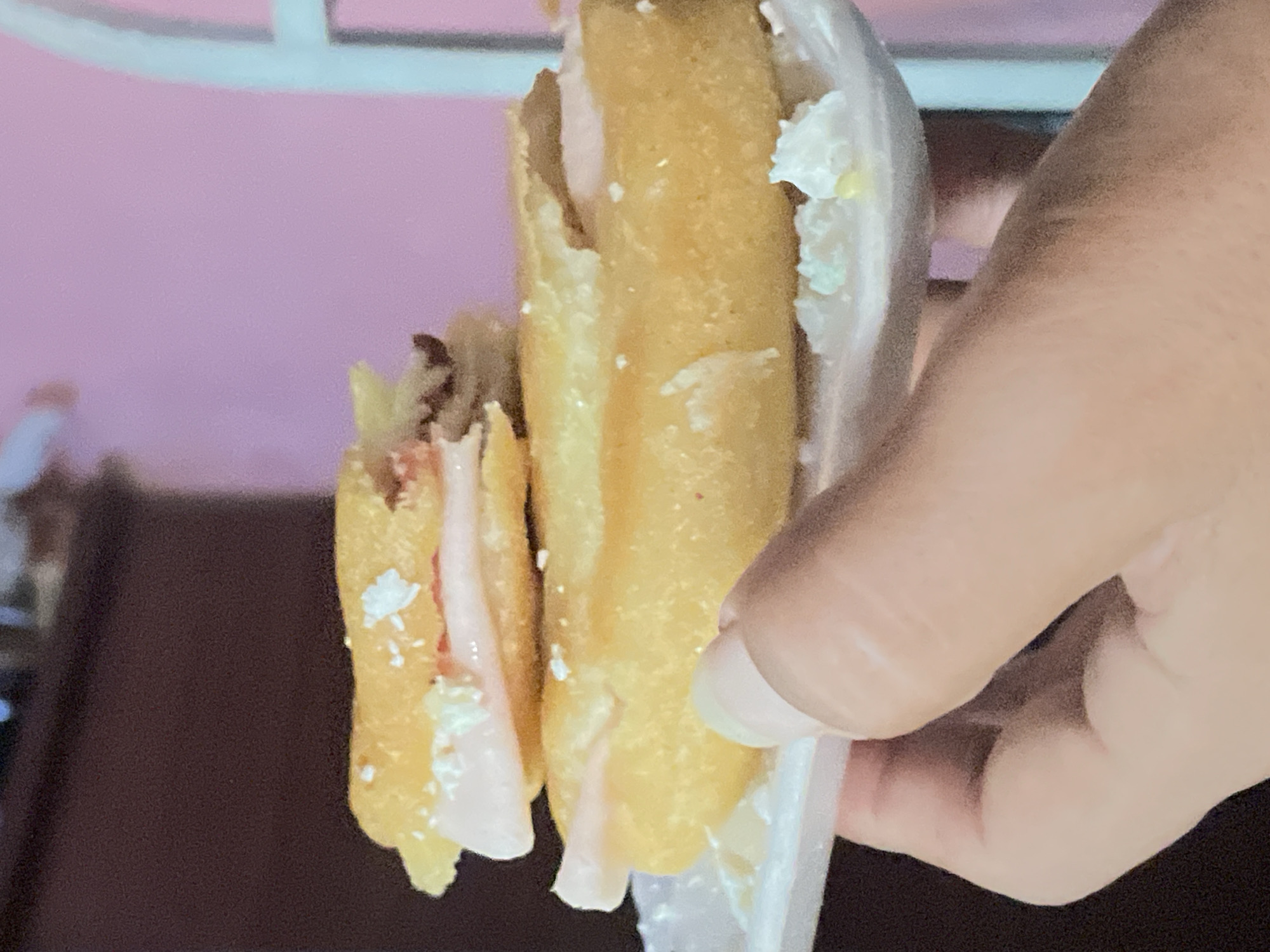 The image depicts two corn dogs held in a plastic wrapper by a human hand. Both corn dogs appear to be partially eaten. The setting suggests an indoor environment with a blurred background of pink and brown hues. The location is identified as Unknown, Unknown.