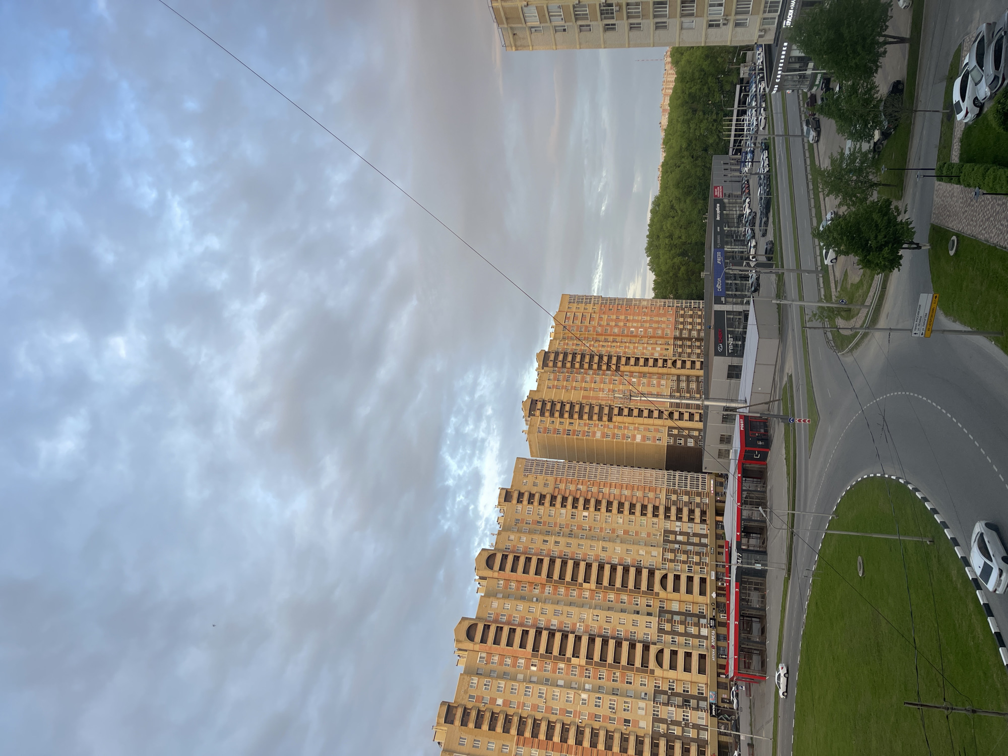 The image depicts a view from an elevated position, likely an apartment building, overlooking a section of Stavropol, Russia. The foreground features multi-story residential buildings with light-colored facades and repetitive window patterns.
Adjacent to the buildings is a roadway with a roundabout, bordered by green grassy areas. Cars are visible on the road and parked nearby. A bus station or terminal with red detailing is also present beside the roadway.
Beyond the roadway, a car dealership or parking area filled with vehicles is visible, lined with trees in the background. The sky is overcast with clouds.