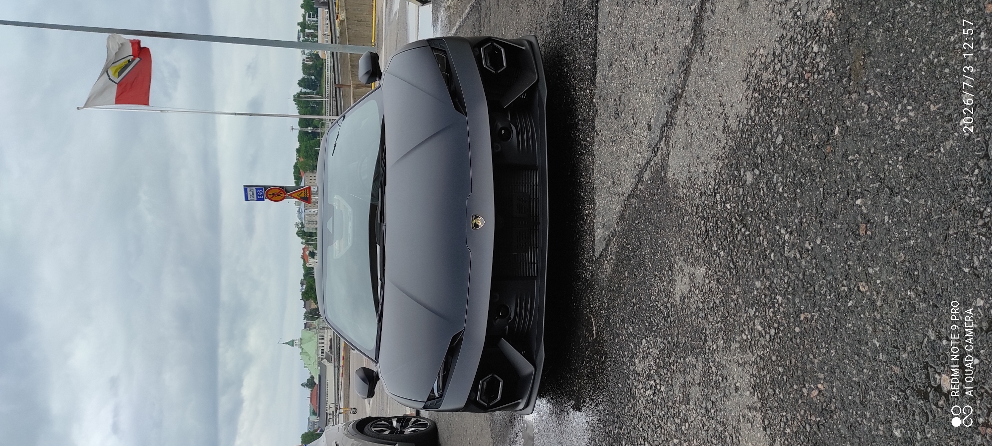 A matte black Lamborghini is parked on a wet, asphalt surface with scattered gravel. The car is facing the camera, with its front grille and headlights prominently displayed. The iconic Lamborghini logo is visible on the hood. In the background, a red and white flag flies from a flagpole. To the left of the car, a portion of another vehicle, possibly an SUV, is visible. Behind the car, there are signs, buildings with green roofs, and a body of water. The sky is overcast and cloudy. The image also contains a timestamp and text identifying the phone model and camera type in the bottom left and bottom right corners respectively.