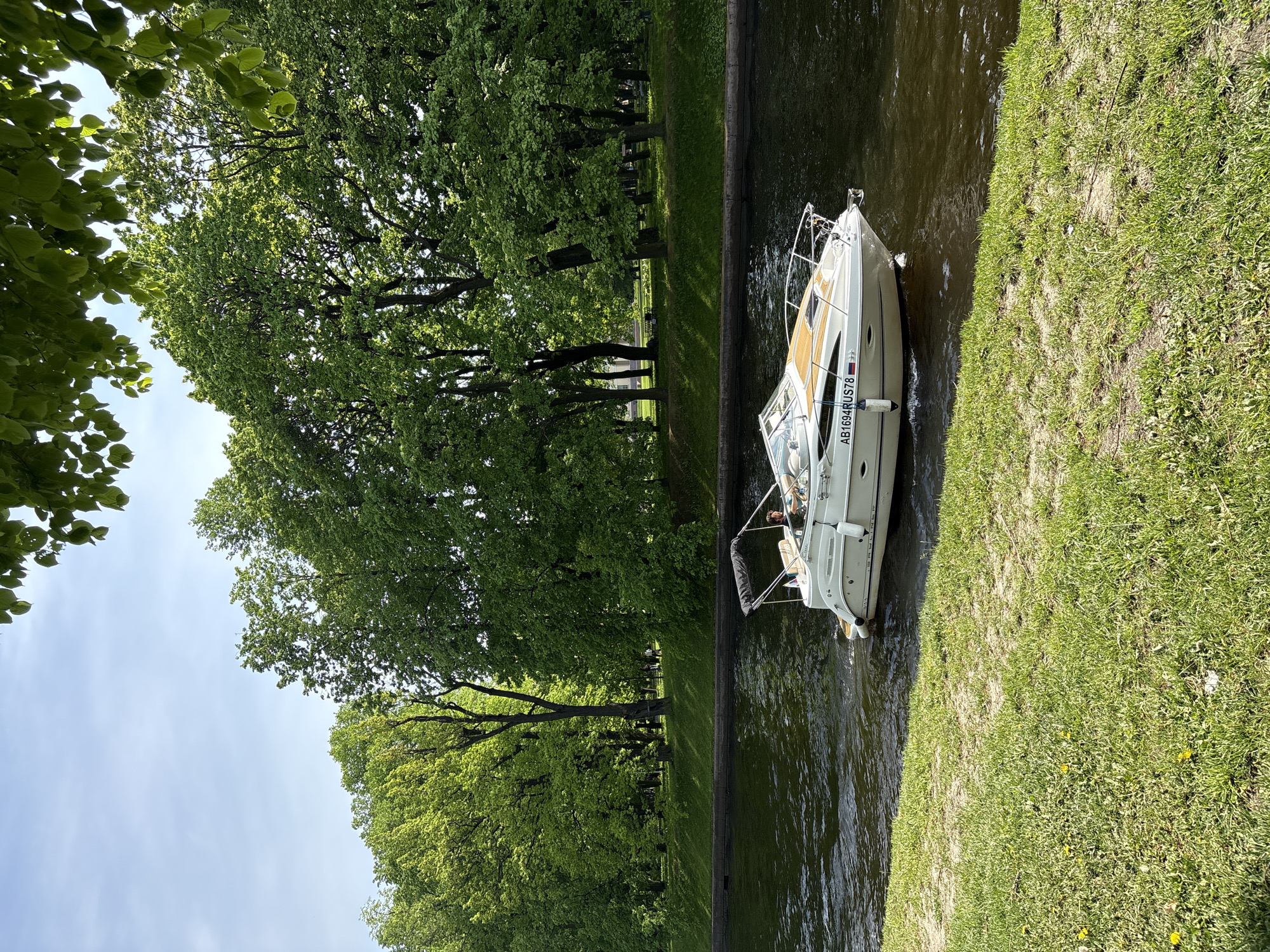 A motorboat is moored in a canal in Saint Petersburg, Russia. The boat is white and has a registration number "AB1694RUS78". The canal is lined with a concrete edge and grass. Trees line the other bank. One person is in the boat.