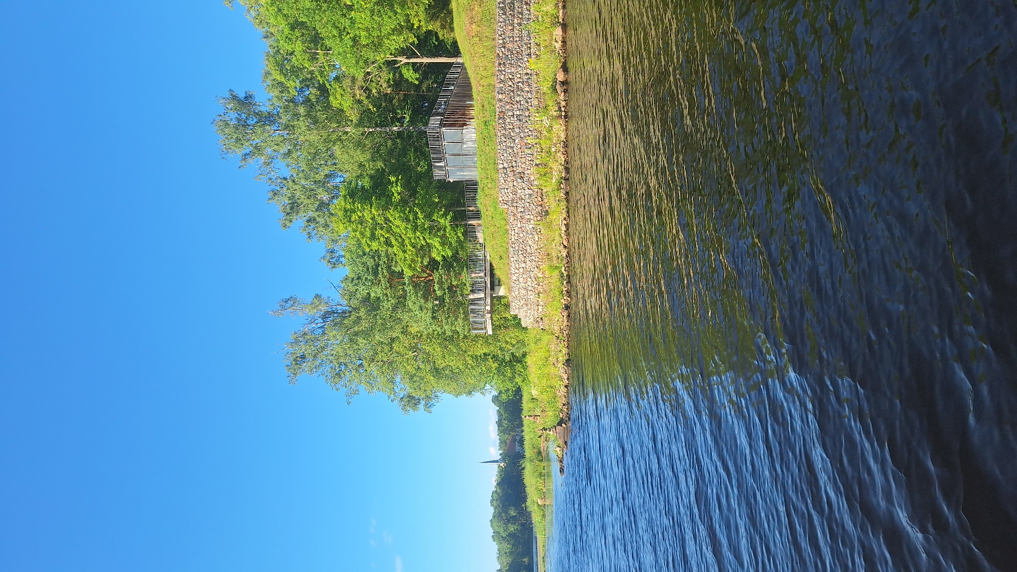 This is a photograph taken from the water, likely from a boat, looking towards the shore in Koknese, Latvia. The foreground is dominated by rippling, dark blue water, reflecting some green from the trees on the bank. On the right side of the image, a stone embankment rises from the water, topped with a grassy slope. At the top of the slope, there is a modern-looking building with a dark facade and large glass windows, accessible by a ramp and a viewing platform with a metal railing. The building is nestled among lush green trees, characteristic of the summer season. To the left of the building, more trees line the shore, and in the distance, across the water and beyond a line of trees, a church spire is visible, suggesting the presence of a settlement. The sky is a clear, bright blue, indicating a sunny day. The overall scene depicts a tranquil waterside landscape with a modern architectural element integrated into the natural environment.