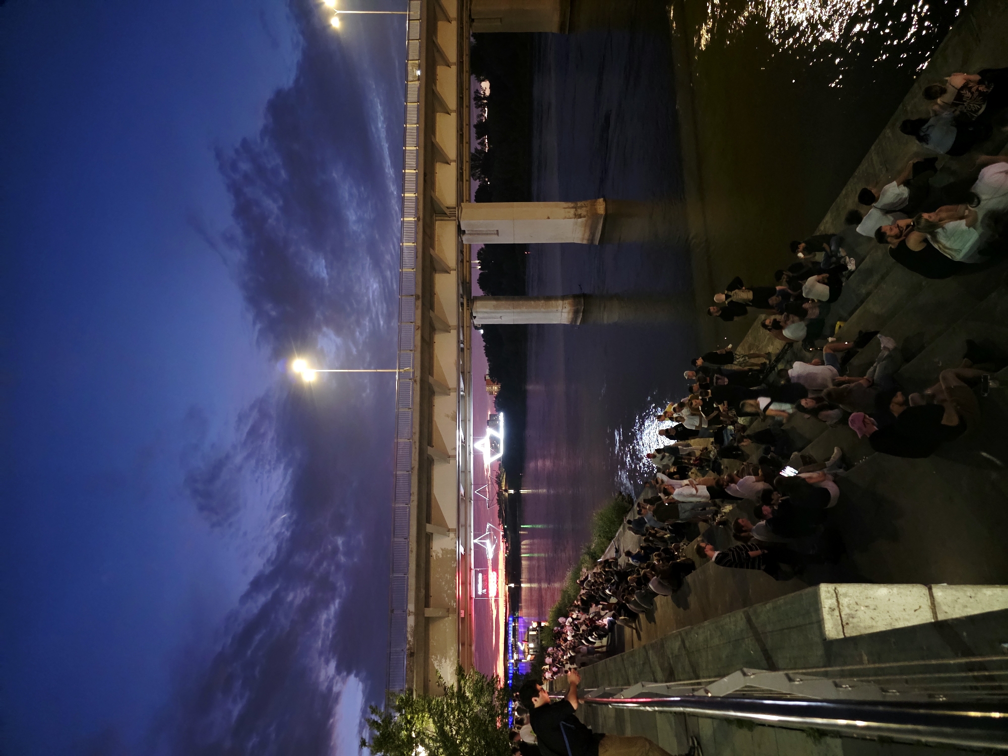 A wide shot captures a riverside scene in Warsaw, Poland, at dusk. A large, concrete bridge spans the river, illuminated by streetlights. Below the bridge, on the riverbank, a crowd of people sits on concrete steps, facing the water. The sky is a deep blue with dramatic clouds, and lights from the city reflect on the water's surface.
The crowd consists of individuals of various ages, most of whom are casually dressed. They appear to be socializing and watching something in the distance, possibly an event or performance on the water or across the river. The atmosphere is relaxed, with groups of people clustered together, some in conversation, while others are looking out at the illuminated cityscape.
The riverbank is lined with concrete structures, including railings and tiered seating areas, indicating a developed urban waterfront. Trees and vegetation are visible on the far bank of the river, and the distant city skyline is dotted with lights. The overall scene suggests a popular public gathering spot where people come to enjoy the evening by the water.