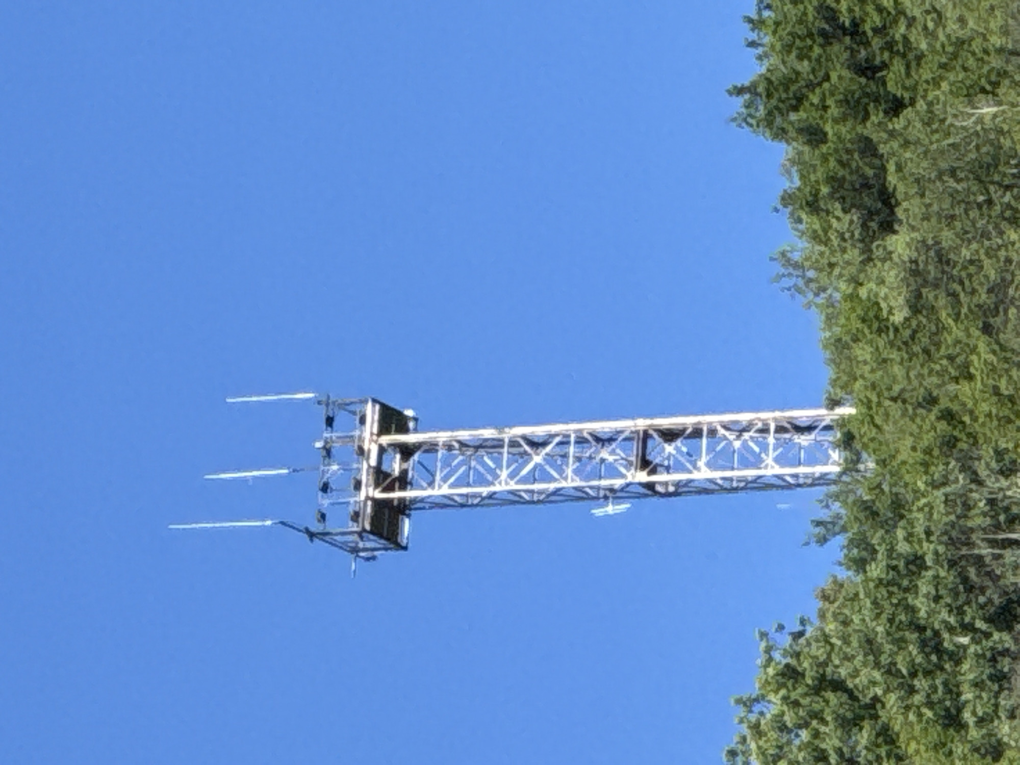 The image shows a lattice tower antenna against a clear blue sky, with a section of tree foliage on the right side. The antenna is a light color, possibly silver or white, and consists of a main supporting structure extending horizontally from a vertical section, which holds several horizontal antenna elements. The vertical section has a square frame appearance with various supporting bars. The tower's lattice structure is composed of interconnected diagonal and vertical beams. The surrounding environment is a blue sky with green foliage in the corner. This antenna could be located in or near Veliko Tarnovo, Bulgaria.