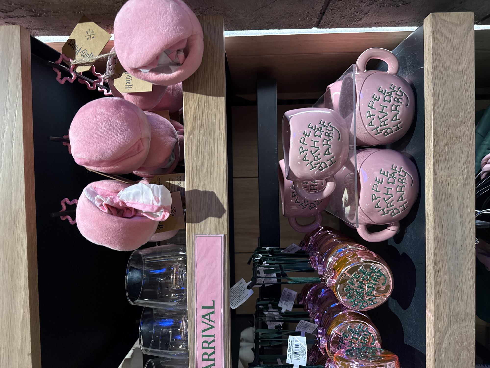 The image shows a display of merchandise, likely in a retail store, featuring pink, plush items and pink mugs. On the left side of the display, there are several round, plush objects, some of which appear to be small bags or pouches. One of these has a white and pink fabric peeking out from its opening. Attached to the plush items are small, brown tags with text that reads "Harry Potter." A pink "ARRIVAL" sign is also visible on this side of the display.
On the right side of the display, several pink mugs with handles are stacked and displayed. Each mug has green lettering that appears to spell out "HAPPEE BIRTH DAE HARRY," celebrating Harry Potter's birthday. Below these mugs, a row of shimmery, pink and gold colored items, possibly cups or accessories, are arranged. Some of these items have green lettering on their bases, which seems to be "TO MAKE YOU HAPPY."
The overall setting appears to be a well-lit indoor retail environment, likely a gift shop or a section of a larger store dedicated to Harry Potter merchandise. The colors and themes suggest a playful and celebratory atmosphere, possibly related to a birthday event or product launch. The presence of price tags with currency symbols and numbers (e.g., "€15.00", "¥2.000") indicates a commercial context. There are no people visible in the image.