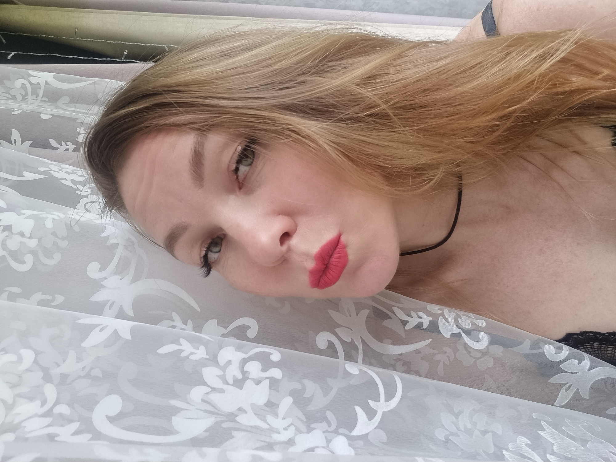 A young woman with long, flowing light brown hair is captured in a close-up portrait, looking directly at the camera. She is lying down or reclining, partially covered by a white, semi-transparent fabric featuring a decorative floral damask pattern in light grey or white. Her lips are pursed into a "kissing" expression, accented by bright red lipstick. Her eyes are light-colored, and she wears prominent eyelashes. A thin black choker necklace is visible around her neck, and a dark strap, possibly from a bra or top, along with a hint of black lace, can be seen near her shoulder. Her skin appears fair with some freckles on her chest area.
The background consists of multiple neatly stacked rolls or bolts of various fabrics and textiles in different muted colors, including grey, beige, olive green, and darker tones. This setting suggests the image was taken indoors, likely within a fabric store, a drapery showroom, or a textile shop, possibly in Chaikovsky, Russia. The overall mood is casual and playful, with the woman posing for the photograph. There is no visible text in the image.