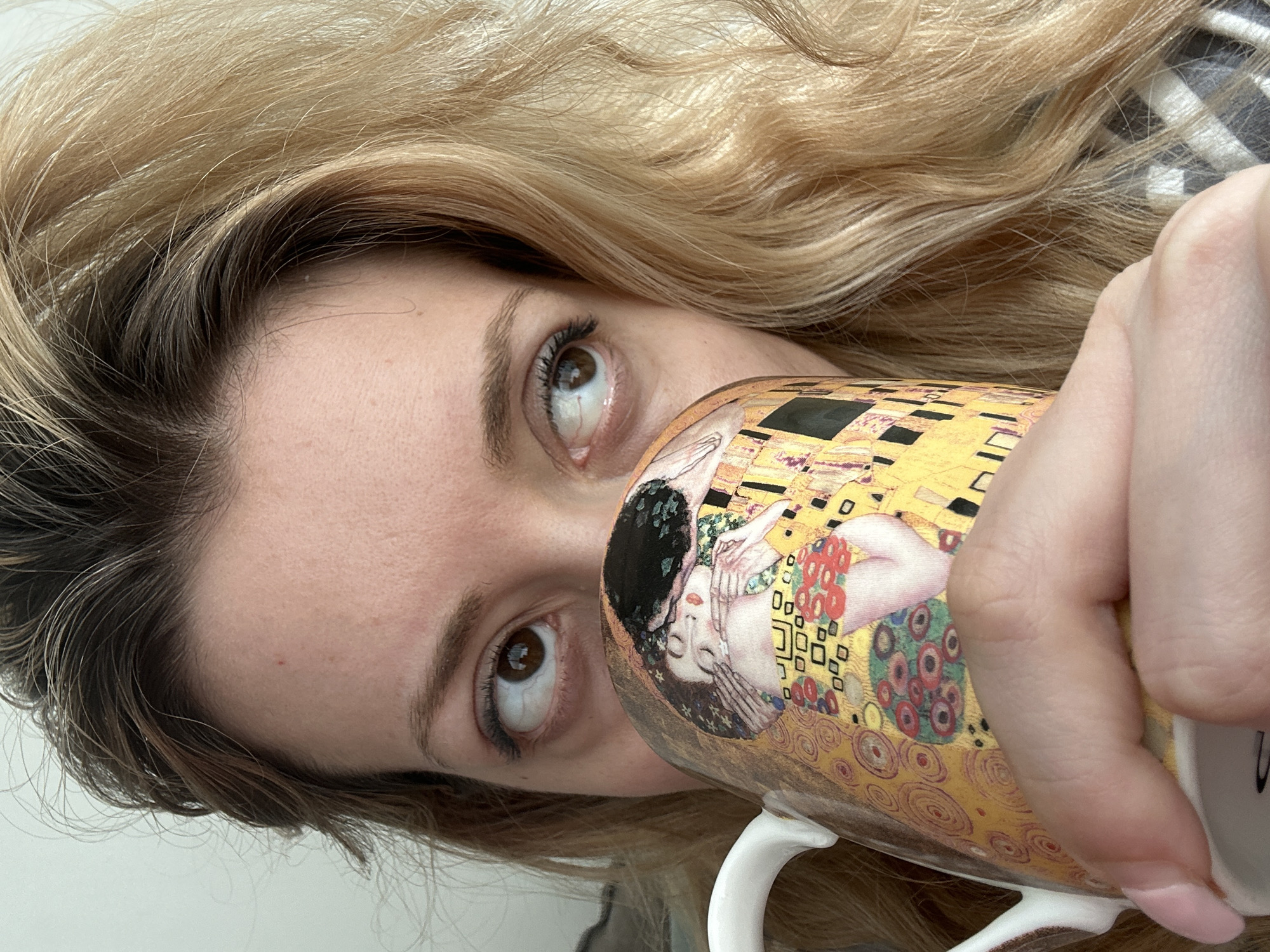 The image presents a close-up, diagonally oriented shot of a person, likely female, holding a mug. The person has long, wavy blonde hair with visible darker brown roots, flowing across the frame. Her face is partially visible, focusing on her right eye and eyebrow, which are looking upwards. The mug, held by her hand, is positioned close to her face, partially obscuring it. The mug is decorated with a detailed reproduction of Gustav Klimt's painting "The Kiss," featuring the embracing couple in rich gold and patterned colors. The person's hand, with manicured nails, wraps around the mug. The background is a plain, light-colored surface, suggesting an indoor setting. There are no explicit location or time-of-day cues. No discernible text is visible on the mug beyond the artwork.