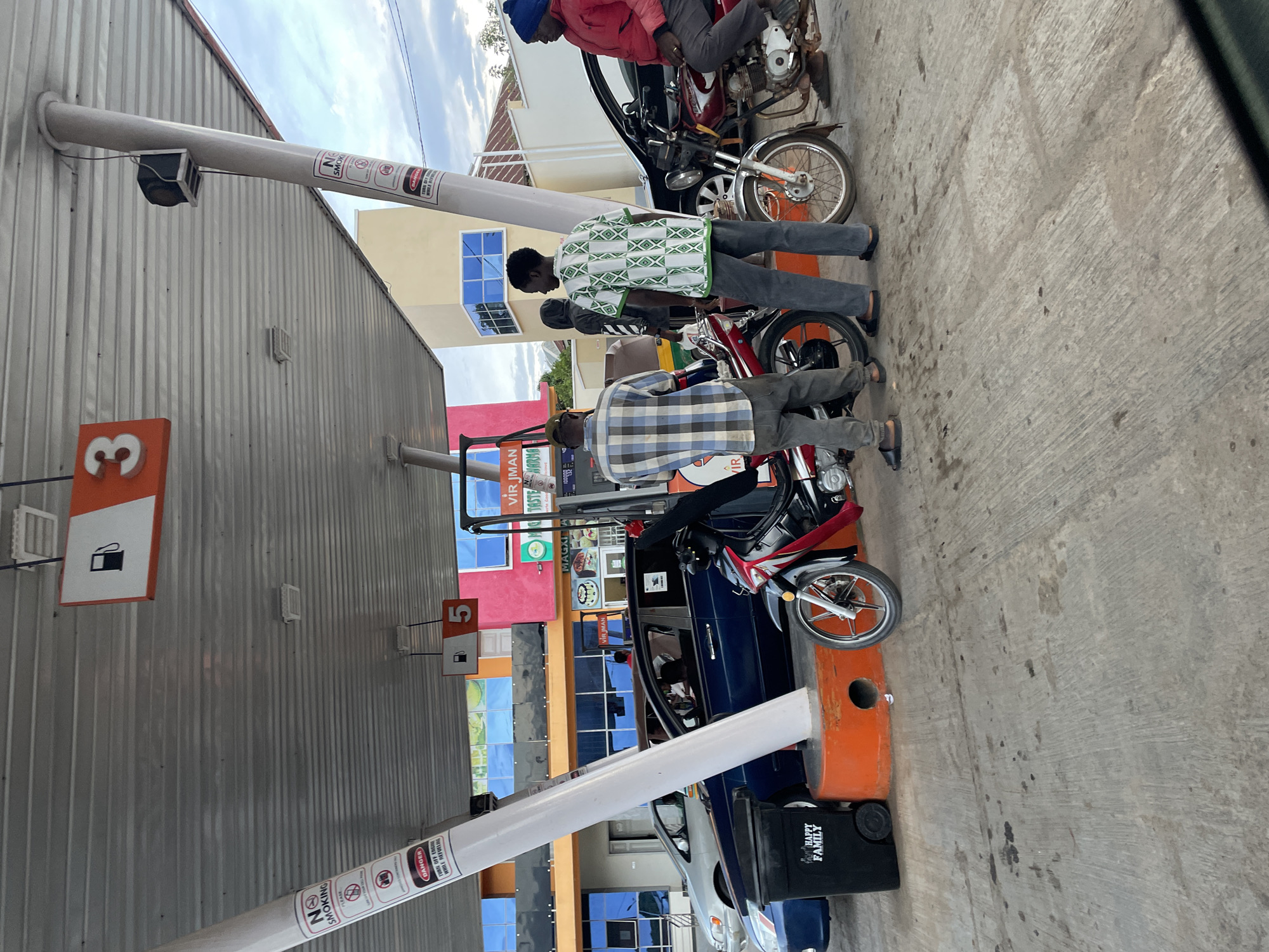 The image shows a scene at a gas station in Jos, Nigeria. Several individuals are standing near motorcycles at the gas pumps, with some appearing to be refueling. There are at least three people visible, wearing casual clothing. The gas station has multiple pumps, marked with numbers and gas icons. The surrounding buildings have colorful facades with windows and signs. There are warnings on the gas pump columns such as "NO SMOKING" and "DANGER: TURN OFF ENGINE WHILE REFUELING." The brand name "VIR JMAN" is visible on signage at the gas station. There are gas tanks and motorbikes. One has the brand name "HAPPY FAMILY."