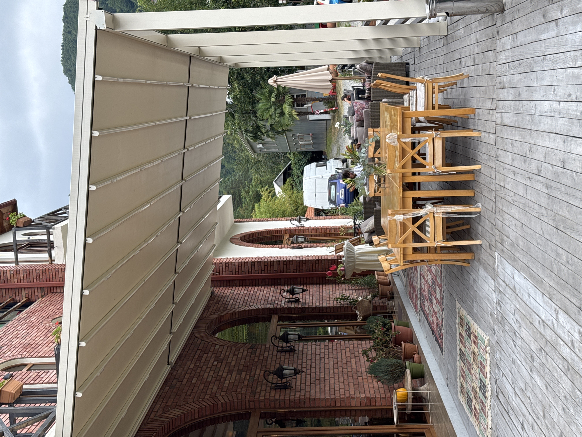 The image shows an outdoor seating area with a retractable awning in front of a brick building with arched windows. A wooden table with chairs is set up on the deck. In the background, there are parked vehicles, including a white van and a blue car, and residential buildings surrounded by greenery. The scene appears to be a cafe or restaurant patio. There are no clear indications of the city of Tibeta, Georgia.
