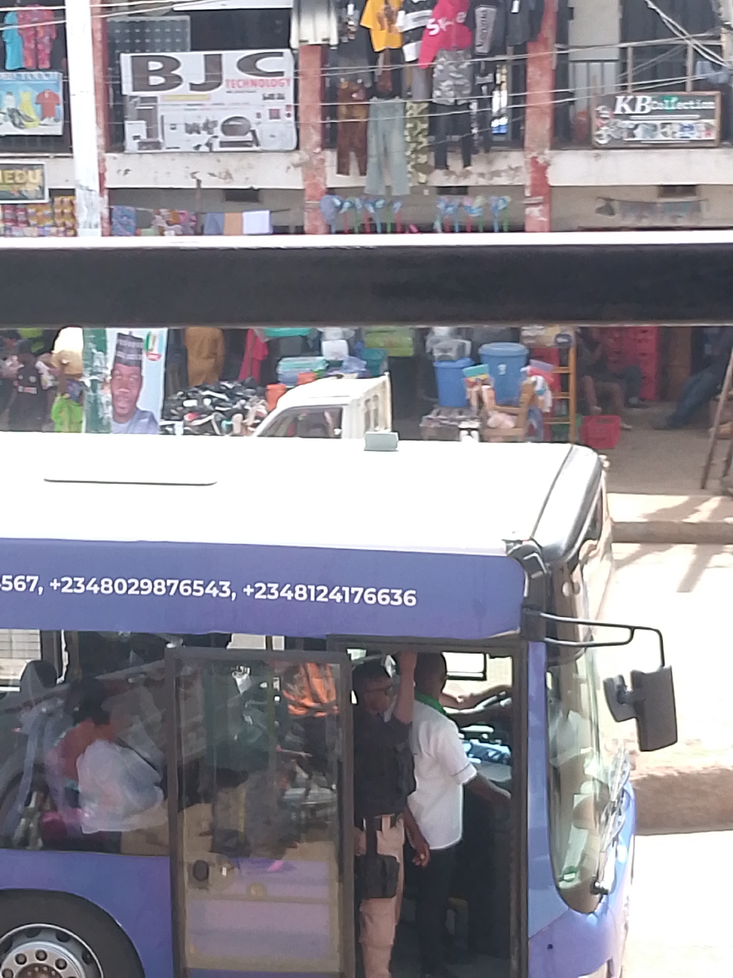 This media file depicts a bustling street scene in Bukuru, Nigeria, likely during the daytime, under bright sunlight. The foreground is dominated by the top and side of a purple bus. The bus has a dark gray roof and a large side mirror. Visible text on the side of the bus reads: "567, +2348029876543, +2348124176636". Through the open door of the bus, several people can be seen inside, some appearing to be passengers and possibly the driver. One man in a white shirt is visible in the driver's seat, and another man in a black shirt stands near the entrance. The interior of the bus appears crowded with luggage.
The background reveals a marketplace with various shops and stalls. Above the street level, buildings display signage. One sign reads "BJC TECHNOLOGY". Another sign is partially visible and says "KBcollection". Clothes are hanging for sale, and numerous plastic containers and crates are stacked, suggesting a commercial environment. The overall impression is one of vibrant activity and commerce. The people appear to be engaged in daily activities, with no strong indicators of specific emotions beyond the general hustle of a market. The setting strongly suggests an urban or semi-urban environment in Nigeria.
