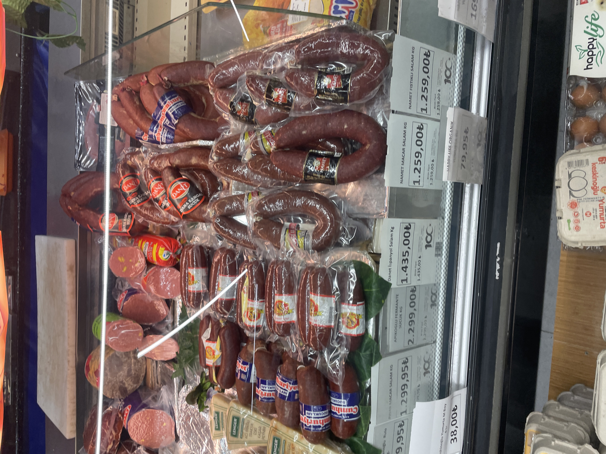 The image shows a refrigerated display case in a Turkish supermarket in Kadıköy, Istanbul. The case is filled with various types of cured meats, primarily sausages, packaged in plastic. There are multiple rows of products, with labels indicating prices in Turkish Lira (₺). Products visible include "Namet Macar Salam Kg," "Namet Fıstıklı Salam Kg," "Danet İspanyol Salam Kg," and "Apikoğlu Fermante Sucuk Kg." In the foreground, there are cartons of eggs and a product labeled "happylife." The lighting is typical of a supermarket, with fluorescent lights illuminating the products. There are no people visible in the image. The environment appears to be a clean and well-organized food retail setting.