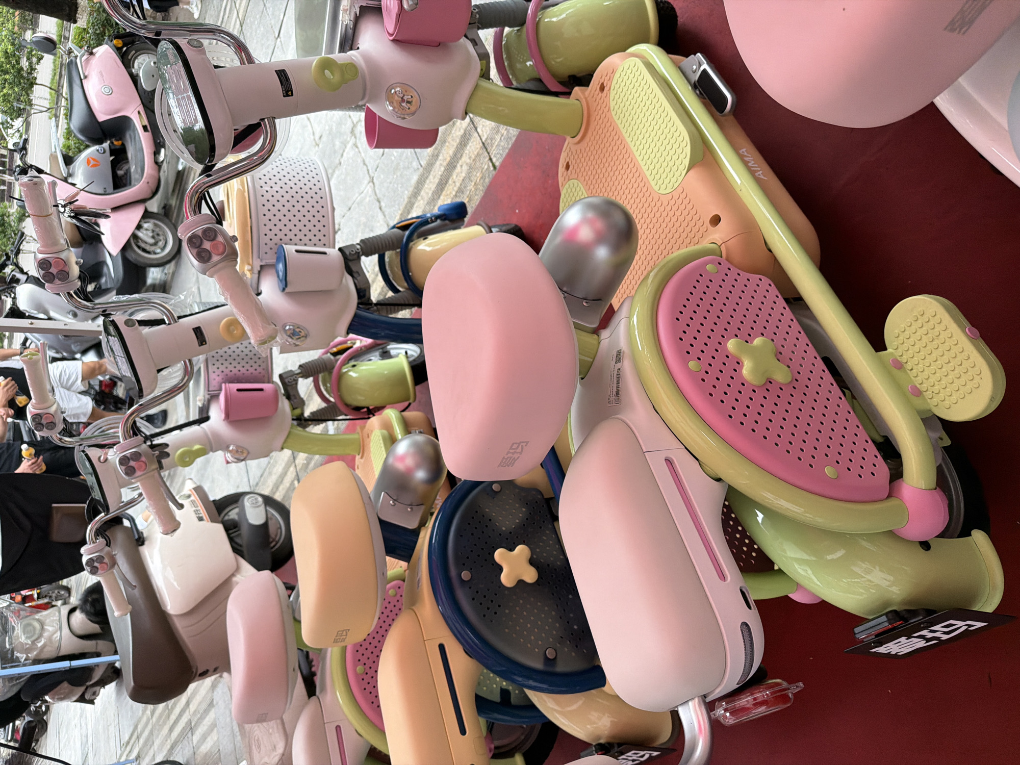 This image shows a chaotic arrangement of pastel-colored children's scooters and electric bikes on a red surface. The scooters are predominantly pink, yellow, and green, with various playful designs. Several electric bikes with similar pastel colors are also visible, some appearing new with plastic coverings.
In the background, blurred figures of people can be seen, suggesting a public space. The location is identified as Chongqing High-tech Zone in China.
The overall impression is one of disarray, possibly from a display or storage area for these children's vehicles.