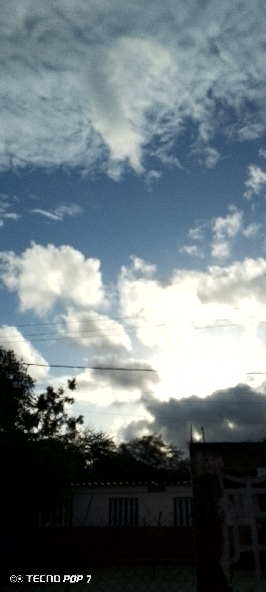 The image captures a view of the sky and rooftops in Altagracia, Venezuela. The sky is filled with a mix of white and gray clouds against a blue background, with the sun partially obscured by the clouds, creating a bright, diffused light. Power lines are visible stretching across the frame. In the lower portion of the image, silhouetted trees and the tops of buildings are visible. One building appears to be a house with windows. The bottom left corner of the image displays a watermark with the text "TECNO POP 7". The overall scene suggests an outdoor, daytime setting.