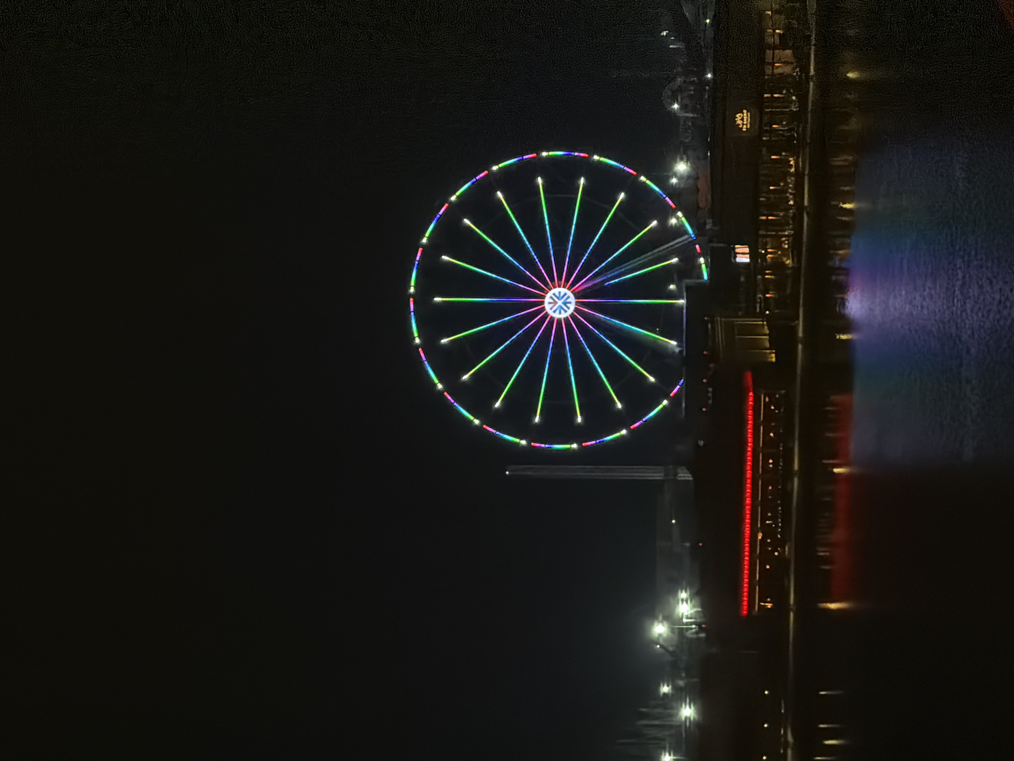 An eye-level, vertical shot captures a brightly lit Ferris wheel against a dark night sky in Unknown, Unknown. The Ferris wheel is the central focus, adorned with a vibrant display of multicolored lights that radiate outward from its center. The spokes of the wheel are illuminated with alternating green, blue, and purple lights. In the background, a long, low-lying building with red lights is visible, reflecting onto the dark water in the foreground. Further in the distance, other structures with illuminated windows and lights are scattered across the landscape. The dark water in the foreground reflects the colors of the Ferris wheel and the surrounding lights, creating a shimmering effect.