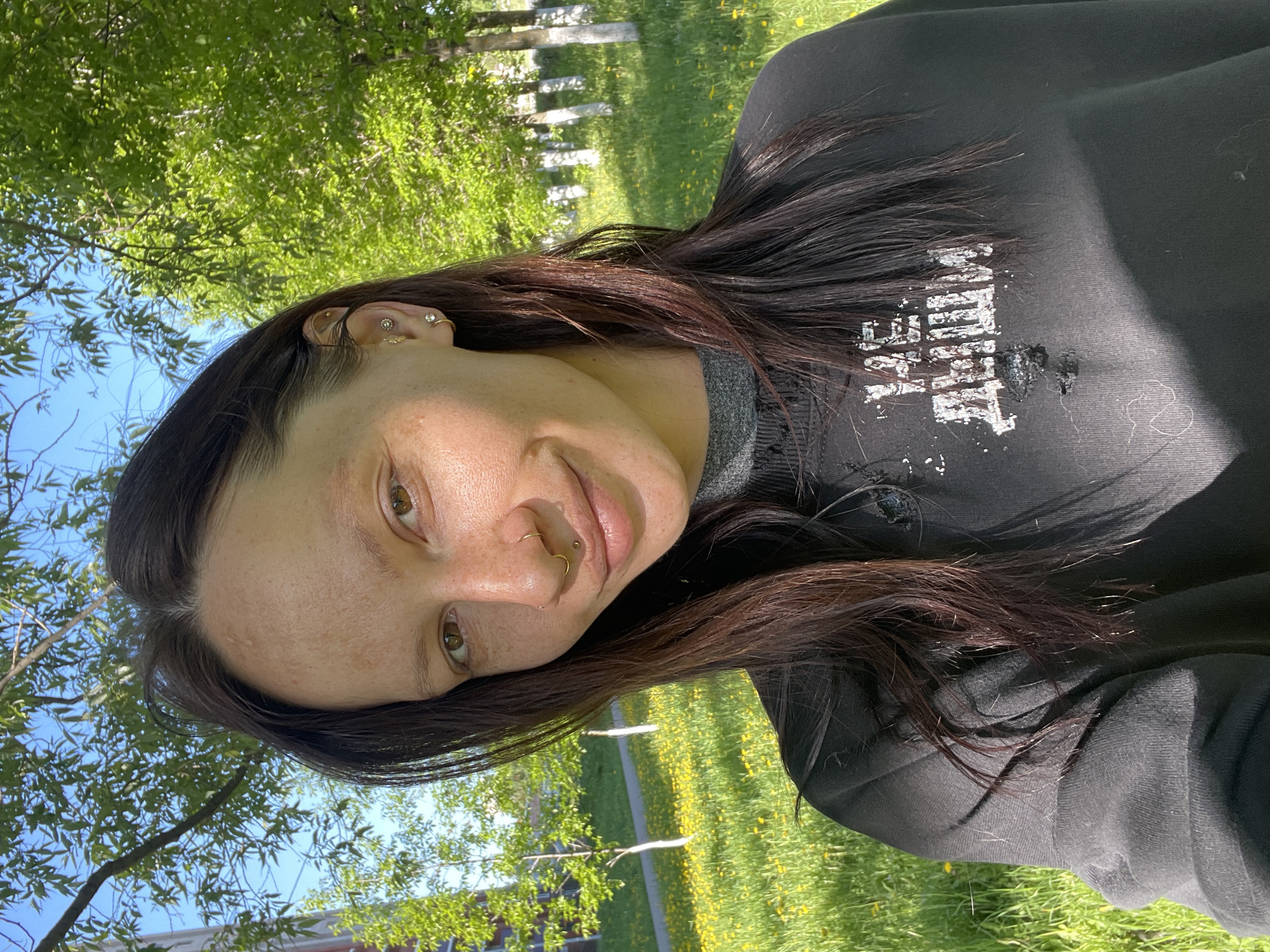 A woman with dark hair and light skin is outdoors in a grassy area. She is wearing a black sweatshirt. She has multiple piercings in her ear and nose. The background includes trees and a field of yellow flowers. A building is visible in the distance. This likely is Izhevsk, Russia, given the query.