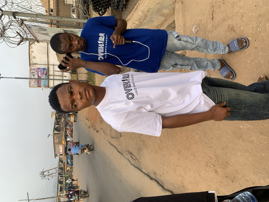 Two men stand side-by-side outdoors. One wears a white t-shirt and jeans, placing his hand in his pocket. The other, behind, wears a blue t-shirt, light jeans, and sandals. He is holding a phone to his ear. Both shirts have a printed logo. The setting appears to be a street scene with buildings and other individuals visible. A street with cracks and some pedestrian traffic suggests an urban environment. Barbed wire and a billboard are visible in the background.