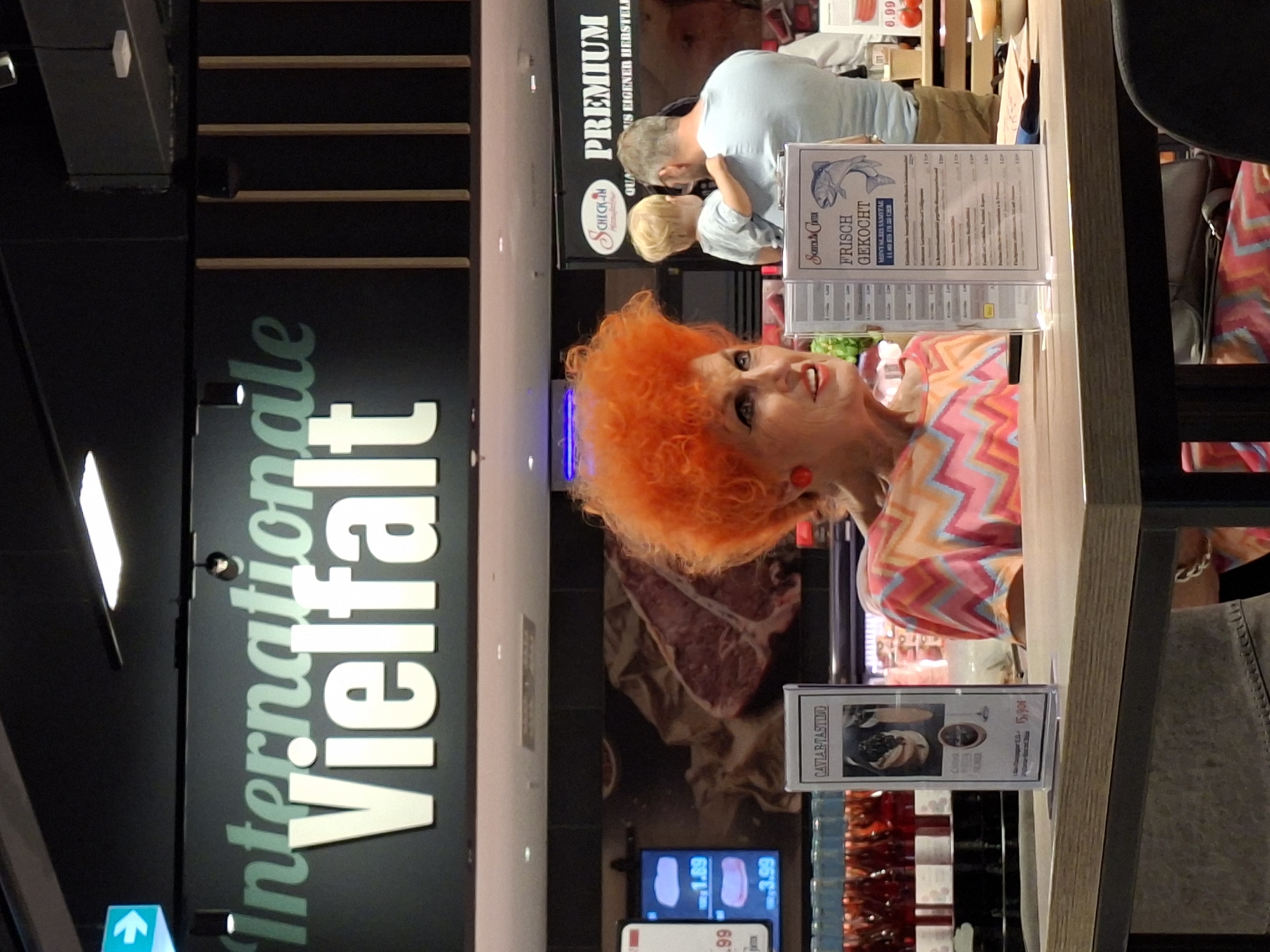 This image appears to be taken inside a store, possibly a supermarket or a specialty food shop, given the signage and products displayed. The primary subjects are two people and various products on shelves.
In the foreground, a woman with vibrant, bright orange, voluminous hair is looking towards the camera with a slightly amused expression. She is wearing a colorful, geometric-patterned top in shades of pink, orange, and yellow. A red earring is visible on her right ear.
Behind her, a man and a child are visible. The man, with graying hair, is leaning towards the child, and they seem to be interacting. The child has blonde hair and is wearing a light blue top.
The background is filled with store shelving and displays. To the left, a large black sign with white and green text reads "Internationale Vielfalt," which translates to "International Variety." This likely indicates a section of the store offering diverse products. Above this sign, there are glimpses of more shelving and what appears to be a ceiling structure.
On a counter or display in the middle ground, there's a sign that says "PREMIUM FRISCH GEKOCHT" (Premium Freshly Cooked), suggesting a prepared food section. Another sign, partially visible, shows what looks like a menu or price list with a fish illustration and text in what appears to be Dutch or German, mentioning "Samstag" (Saturday).
Several digital price displays are visible, showing prices like "1.89," "3.99," and "15,90€" next to product images. One product advertised is "CAVIAR TASTINO." There are also rows of bottled beverages, some appearing to be red wine.
The lighting suggests it's indoors, likely during the day or under artificial store lighting. The overall atmosphere seems casual and perhaps a bit quirky due to the woman's distinctive hairstyle.