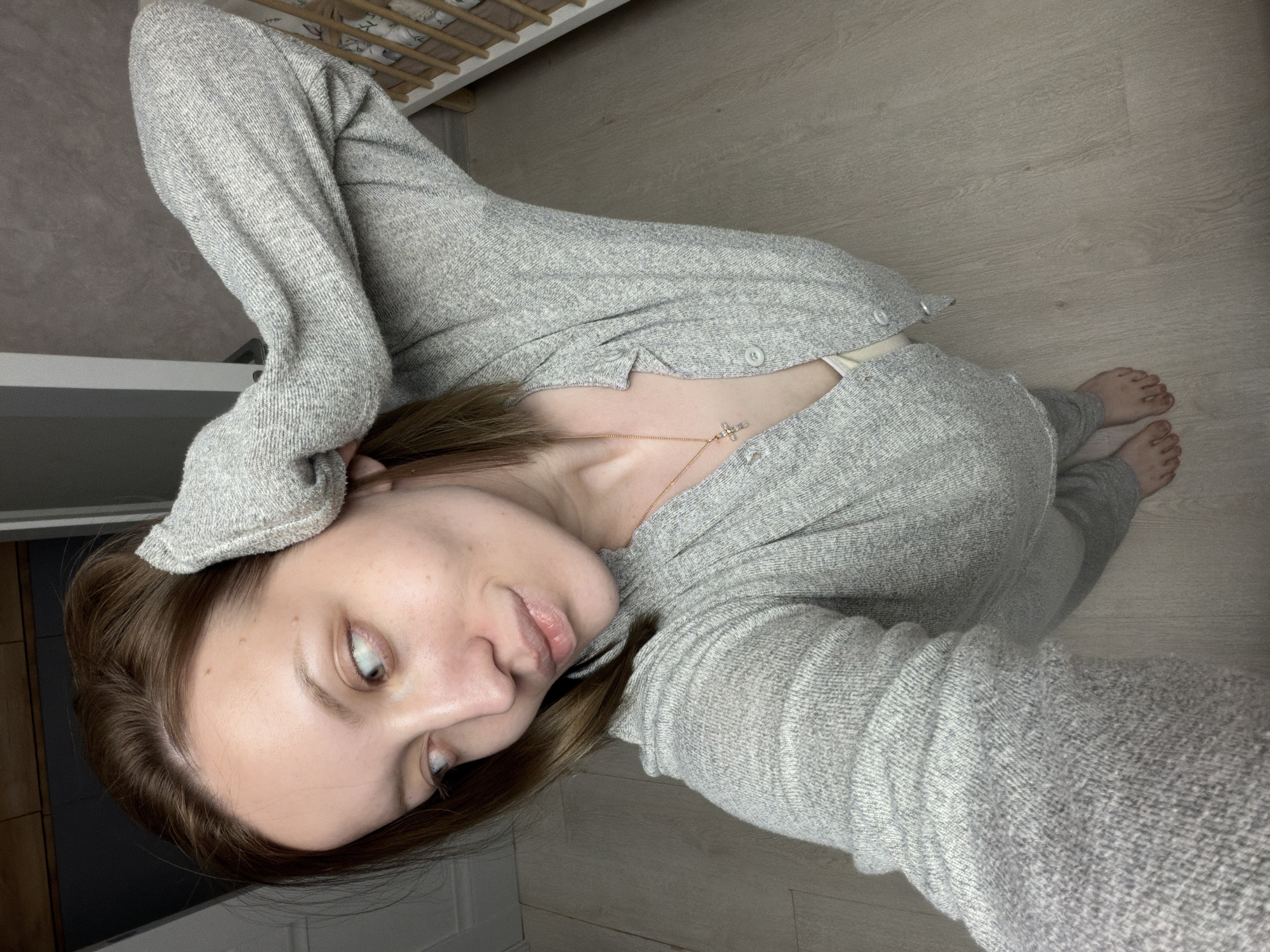 A person is lying on a light-colored wooden floor, wearing a gray knit cardigan and pants. A necklace with a small pendant is visible on their chest. Part of a light-colored crib is visible in the background. The location is an interior space in what is possibly a domestic setting. The location in the city of Reshetnikovo, Russia cannot be determined from the image.