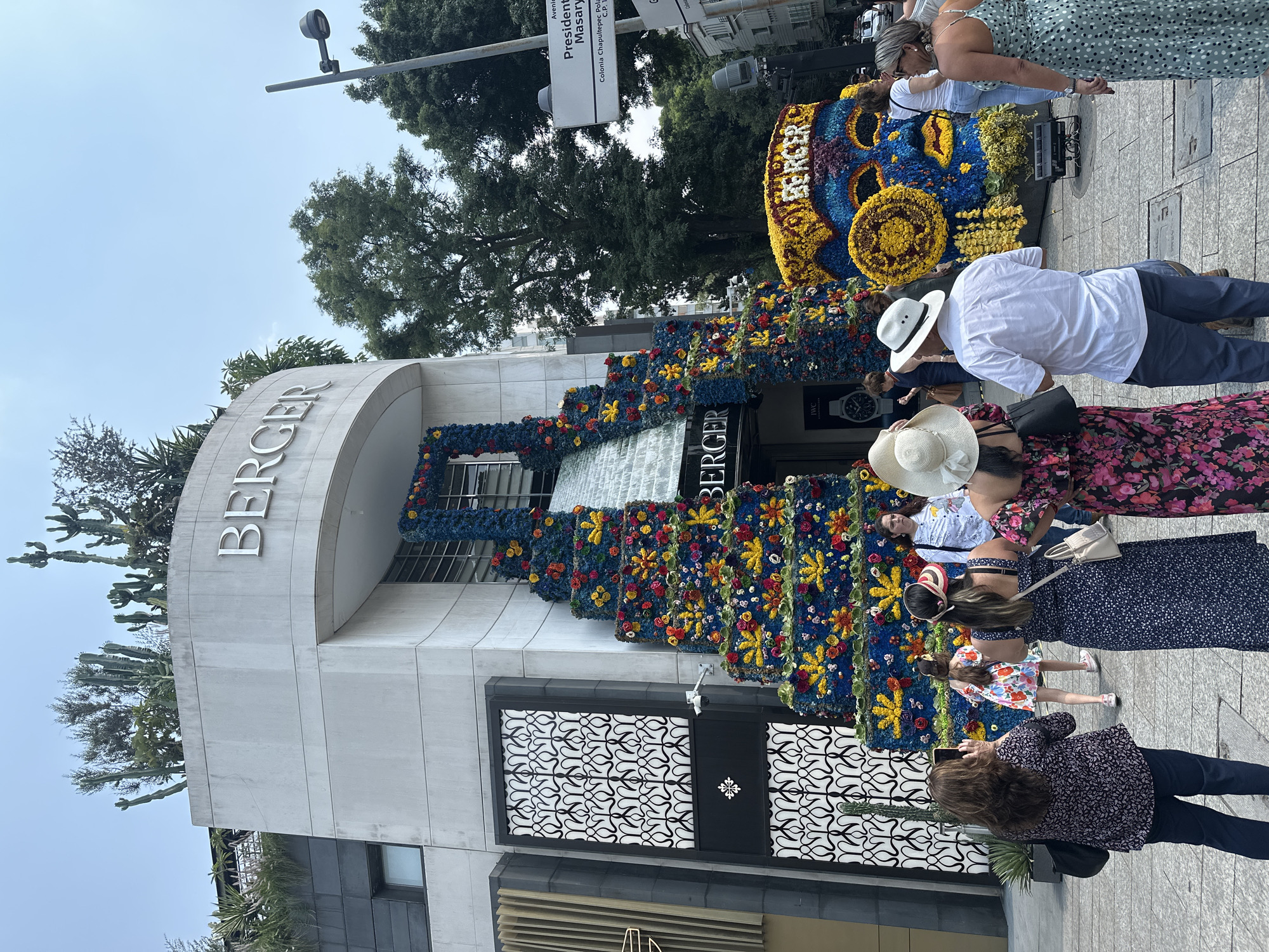 People are standing in front of the Berger Jewelry store in Mexico City, Mexico. The building is decorated with an elaborate floral installation that spills down the front. Pedestrians are admiring the display. The street sign "Presidente Masaryk" is visible above the people.
