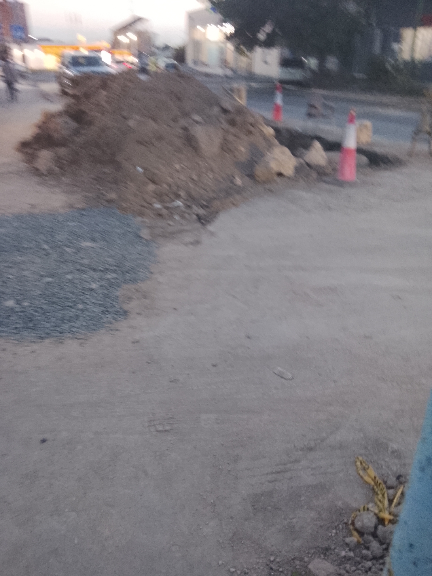 A pile of dirt and gravel sits in the foreground of an outdoor scene. Two orange and white traffic cones are visible to the right of the dirt pile, standing on a paved surface. In the background, vehicles and buildings can be seen, some of which are illuminated. The overall lighting suggests it is either dawn or dusk.