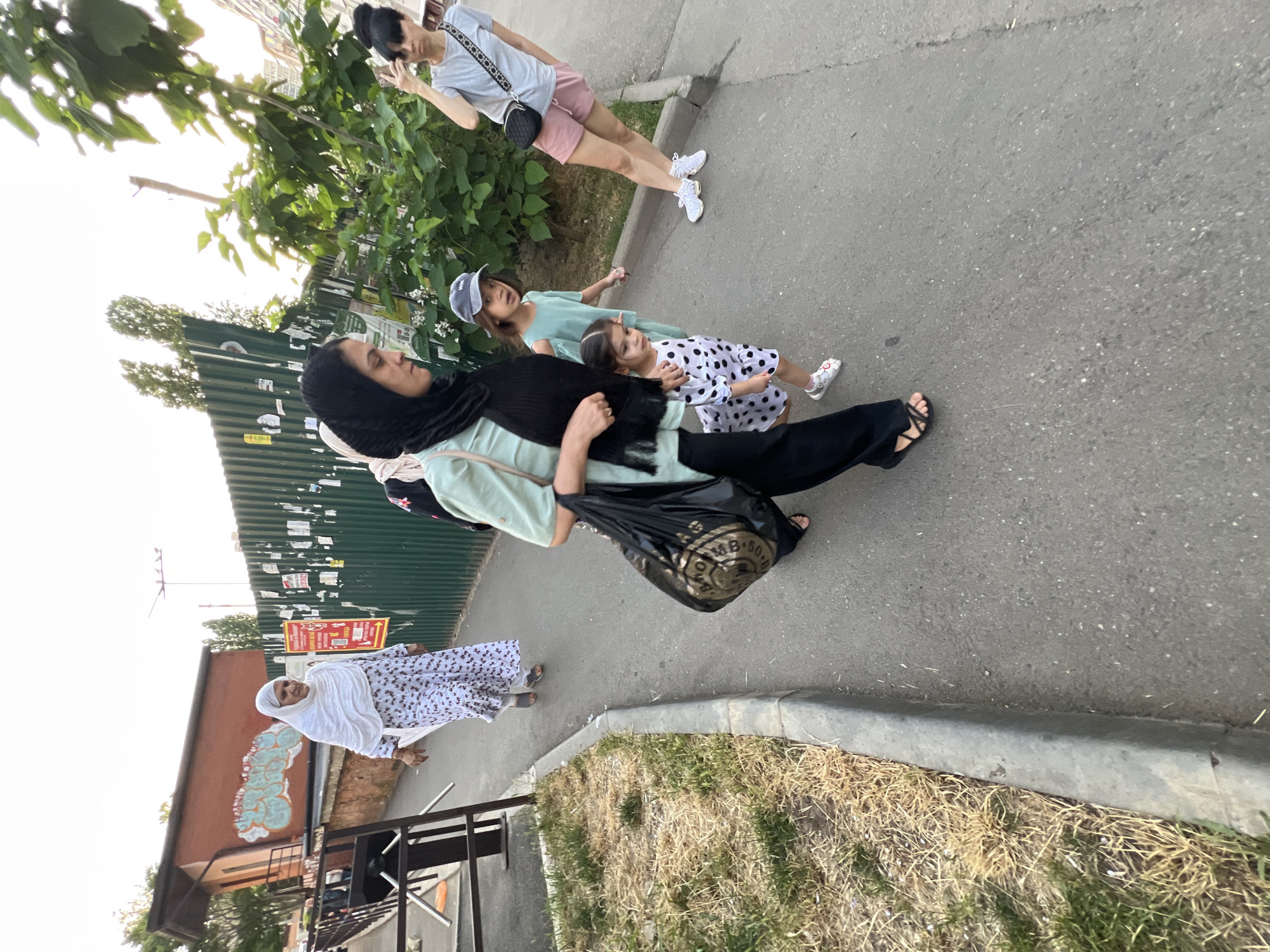 In Krasnodar, Russia, an outdoor scene captures a group of people walking along a paved pathway. The image features an elderly woman in a white hijab and a long, patterned dress, walking away from the camera. To her right, a younger woman in a black hijab and a light green top, also walking away, carries a large black shopping bag. She is accompanied by two young girls. One girl, wearing a light green dress and a blue baseball cap, walks beside the woman. A smaller girl in a black and white polka dot dress walks slightly ahead and to the left of the woman, looking towards the camera. Further to the right and slightly behind, a woman in a grey t-shirt and pink shorts is captured from the waist down, holding a phone to her ear as if taking a photograph. The pathway is bordered by dry, yellowed grass on the left and a green metal fence on the right. A portion of a brick building with graffiti is visible on the far left. The sky is overcast and the lighting suggests it is late afternoon or early evening.