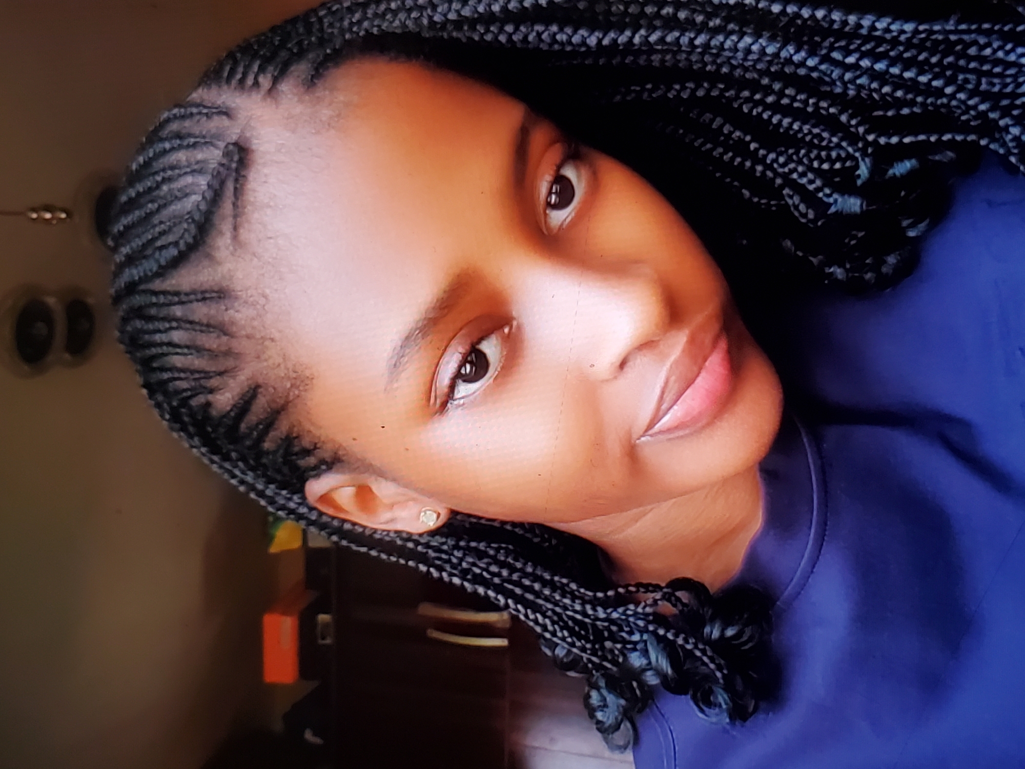 The image shows a close-up, waist-up portrait of a young Black woman with braided hair. She is looking towards the camera with a slight smile. She is wearing a dark blue shirt. The background is blurry and appears to be indoors, with some shelving and objects visible on them. The lighting suggests it is daytime, with light coming from an unseen source. There are no contextual clues to determine the specific location or city.